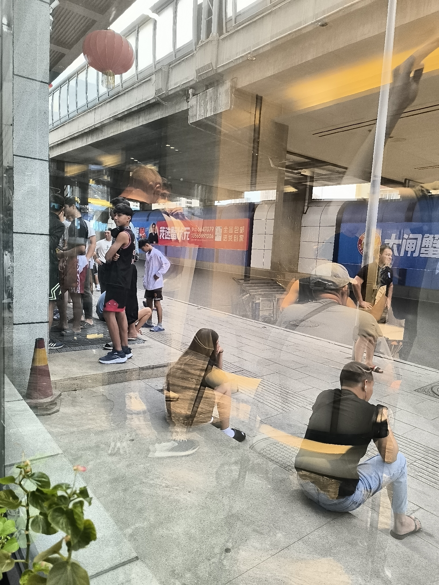 The image shows a reflection on a window, depicting a street scene in Ürümqi, China. Several people are visible, some standing and some sitting on the pavement. There are advertisements on a building in the background. A red lantern hangs from the ceiling above. The scene appears to be an outdoor public area, possibly a bus stop or waiting area, with a pedestrian walkway and a road or platform visible in the reflection. The lighting suggests it is daytime.