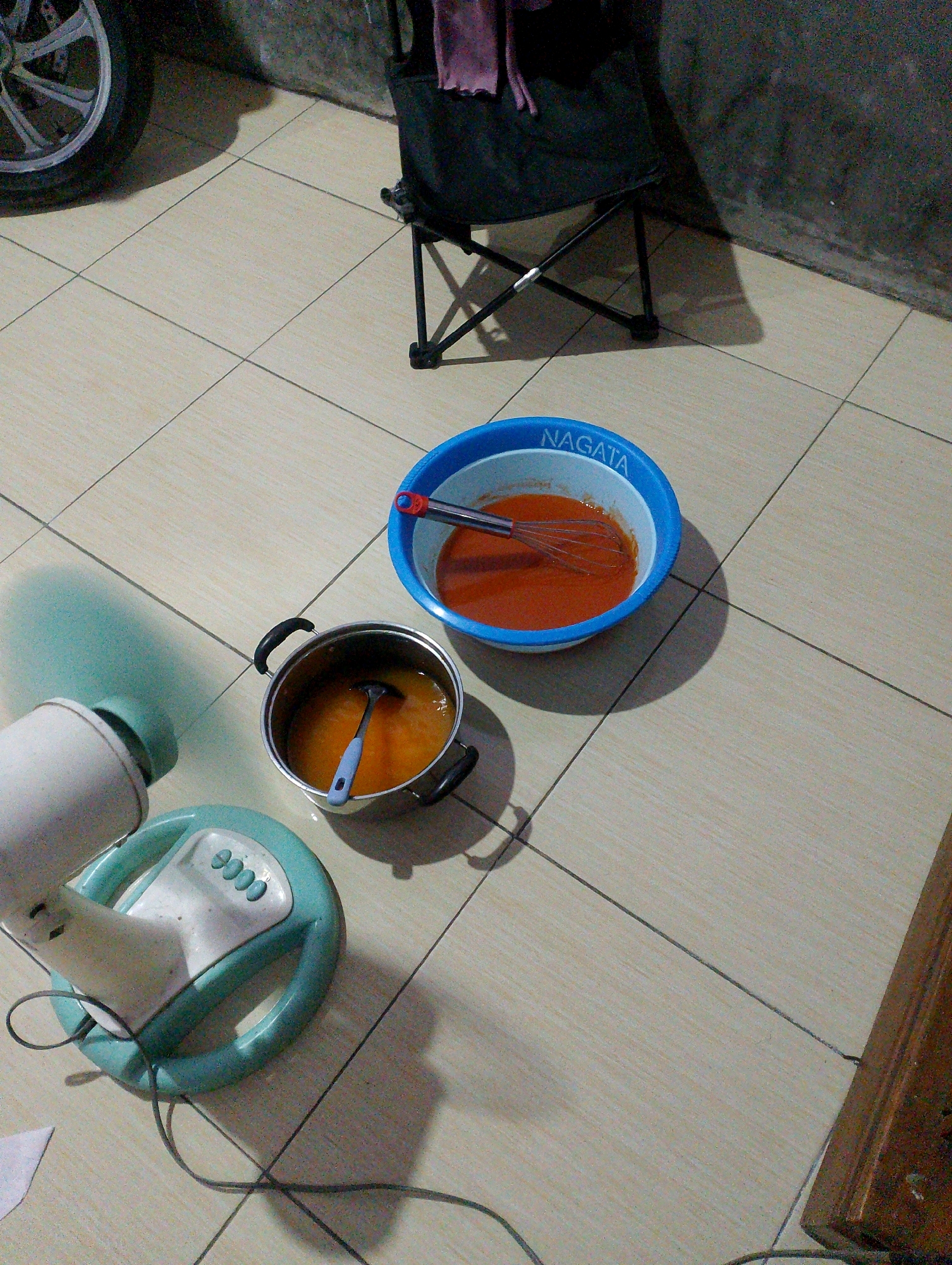 This is a medium-angle, full-shot, eye-level, indoor photograph taken in Desa Babakan, Indonesia. The scene is a tiled floor with a collection of domestic items. In the lower left, a white and teal-colored electric fan with a circular base is visible, its power cord trailing across the tiles. To the right of the fan, a silver pot with black handles contains an orange liquid and a blue ladle. Further to the right, a large blue bowl labeled "NAGATA" holds a reddish-orange liquid and a whisk. In the background, a dark camping chair with a pink cloth draped over it casts a shadow. A portion of a silver car wheel is also visible in the upper left corner. The overall lighting suggests an indoor environment, possibly at dusk or under artificial light.