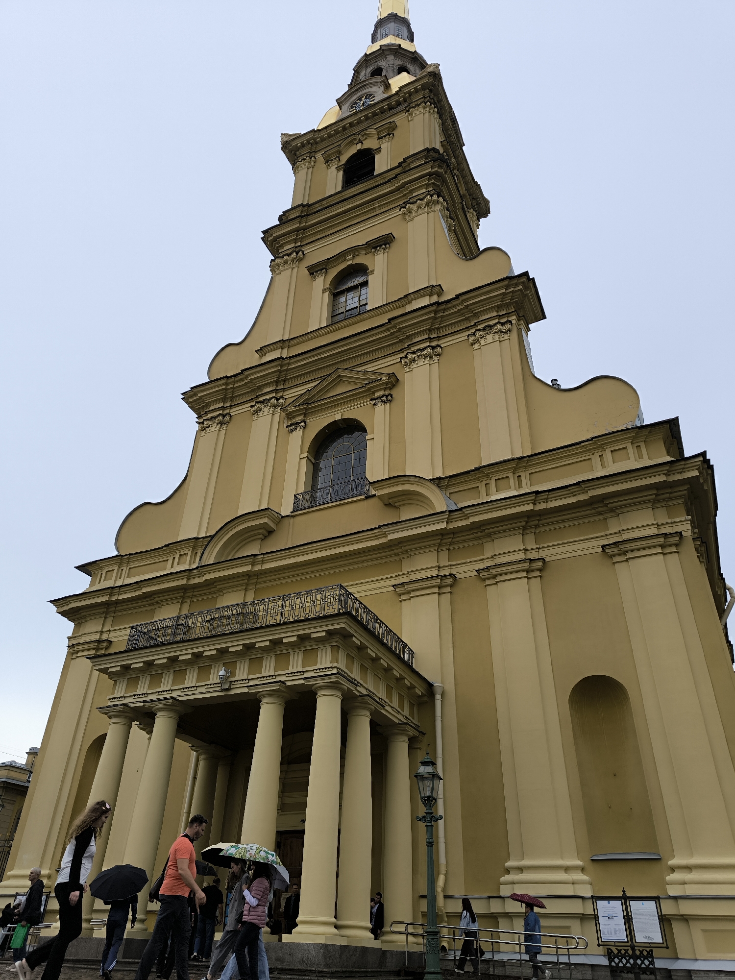 The image shows the Peter and Paul Cathedral in Saint Petersburg, Russia. The cathedral is a tall, yellow building with a gilded spire. The photograph is taken from a low angle, looking up at the cathedral. In the foreground, there are several people walking with umbrellas, suggesting it is raining or has recently rained. The sky is overcast.