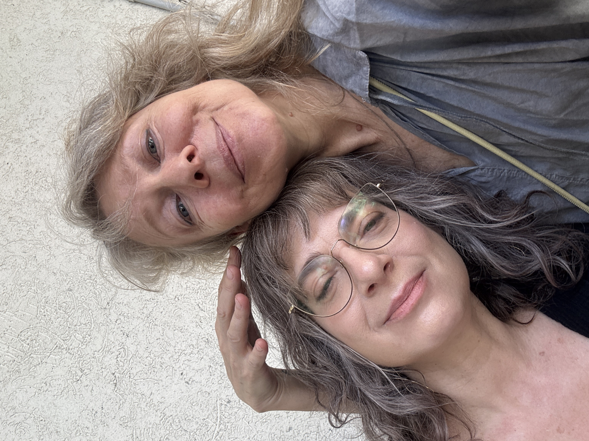 Two women are posing for a close-up selfie. The woman on top has blonde and graying hair and is wearing a gray shirt with a yellow zipper. The woman on the bottom has graying wavy hair and is wearing round eyeglasses. Her hand is near her face. The background appears to be a textured off-white wall. It is not possible to confirm the specific location in Givatayim, Israel, from the image.