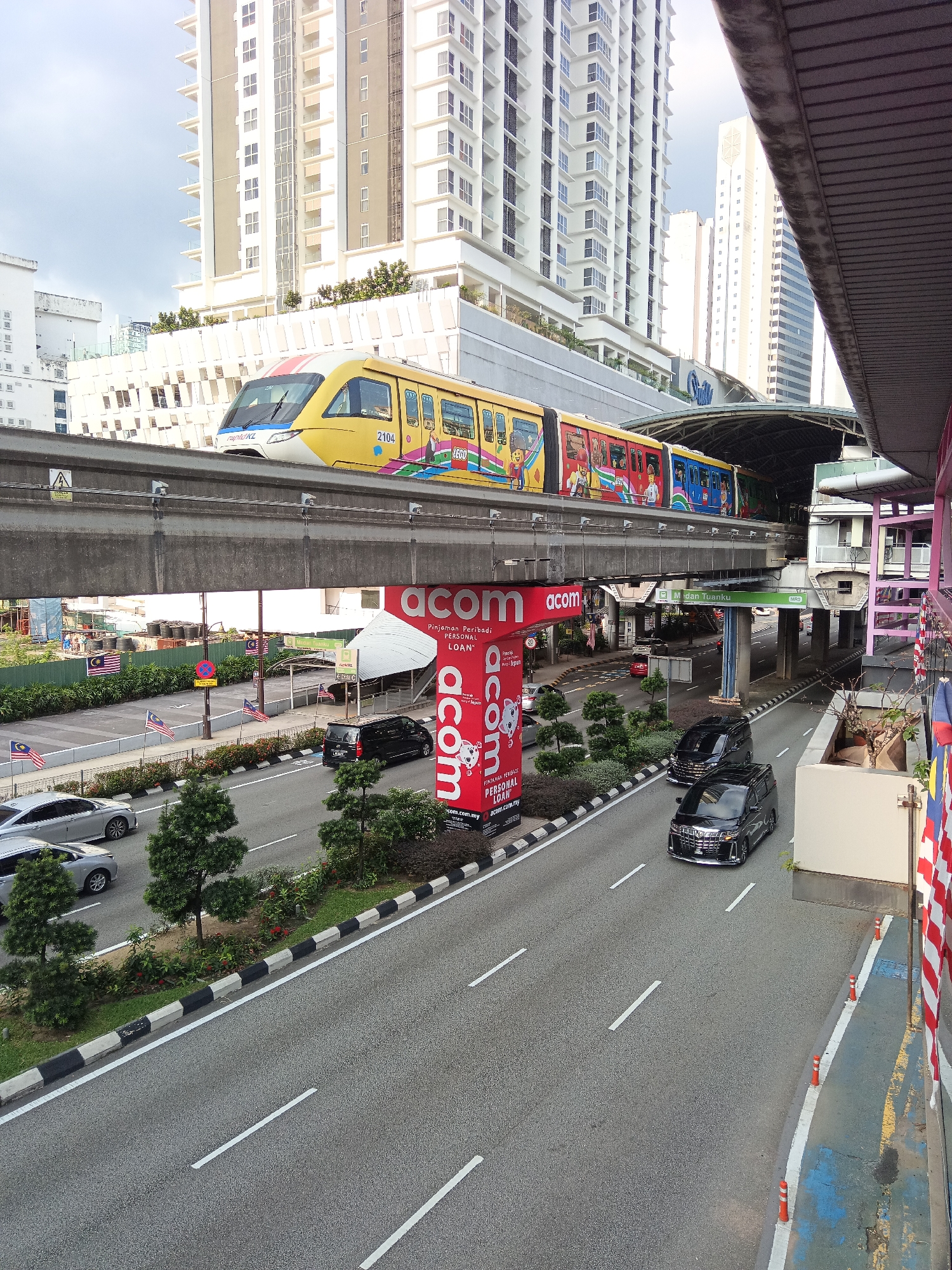 The image depicts a street scene in Kuala Lumpur, Malaysia, featuring a monorail train passing overhead on an elevated track. The train is yellow with colorful cartoon graphics and the number "2104" visible. Below the monorail track, a multi-lane road runs horizontally across the frame, with several cars, including black minivans and silver sedans, driving on it.
Adjacent to the road is a pedestrian walkway and a landscaped area with trees and bushes. A large red billboard with the "acom" logo and the text "Pinjaman Peribadi PERSONAL LOAN" is prominently displayed on the right side of the image. Behind the monorail track and the billboard, modern high-rise buildings with many windows dominate the background, characteristic of Kuala Lumpur's urban landscape. The sky is overcast. Malaysian flags are visible on poles along the roadside. The scene appears to be a busy urban intersection.