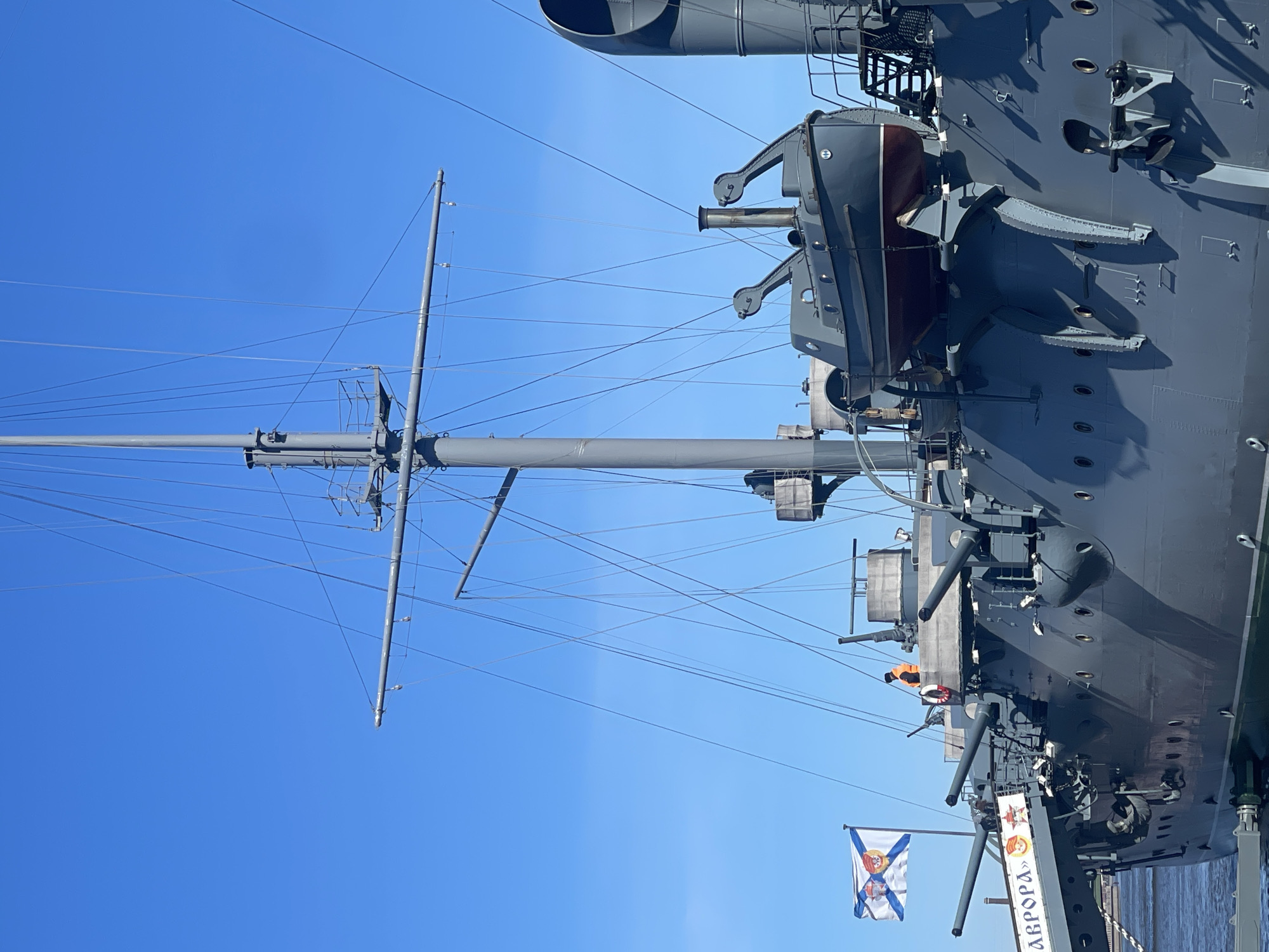 A low-angle, horizontal view showcases the Cruiser Aurora, a historic Russian warship, set against a clear blue sky. The vessel, painted in shades of gray, is docked at the Petrograd Embankment in Saint Petersburg, Russia. A tall mast with various lines and antennas extends upwards from the ship. Prominent features include gun turrets, a lifeboat suspended from davits, and other deck structures. A naval flag is visible near the base of the ship, next to a sign with Russian Cyrillic script that spells "Aurora" (АВРОРА). A person wearing an orange vest is partially visible, positioned on the deck near a gun turret. The ship's hull faces the viewer, showing the multiple portholes along its side.