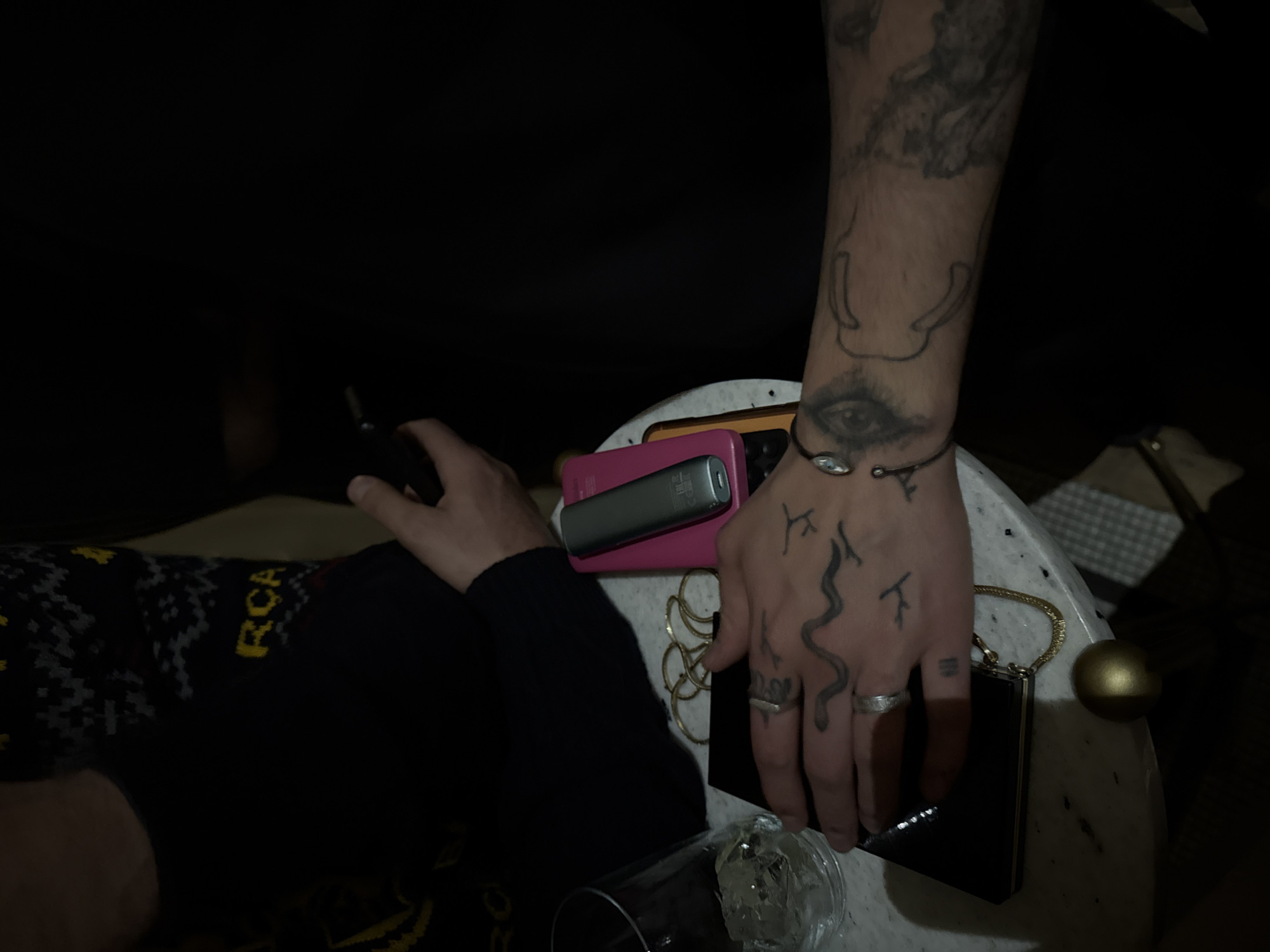 The image depicts a dimly lit indoor scene. A person with arm and hand tattoos, including a stylized eye and a snake, is resting their tattooed arm on a small, round marble table. On the table, there is a pink cell phone, a silver vaping device, several gold bracelets, a small black purse with a gold handle, and a partially visible glass of water with ice. Another person, wearing a dark sweater with yellow lettering that reads "RCA", is visible to the left, holding a small, dark object in their hand. The location of Yerevan, Armenia, cannot be confirmed from the image.