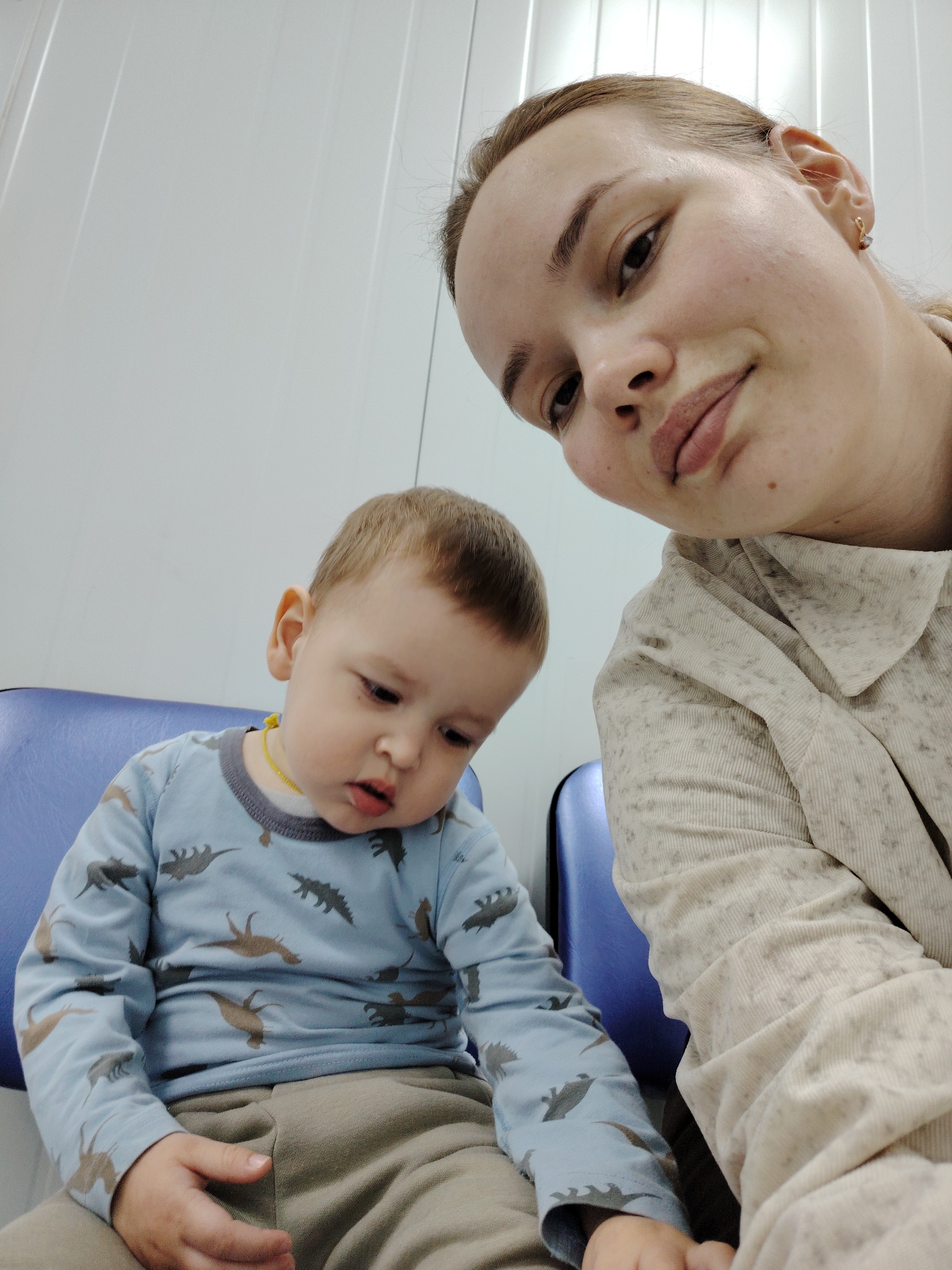 A woman and a young child are sitting next to each other. The woman is holding the camera at a high angle, taking a selfie. The child is sitting on a blue chair wearing a blue, dinosaur-patterned shirt and khaki pants. The woman is wearing a button-down shirt with a textured pattern. The background is a plain white wall. The location is likely a waiting room or a similar clinical setting in Марковский, Russia.