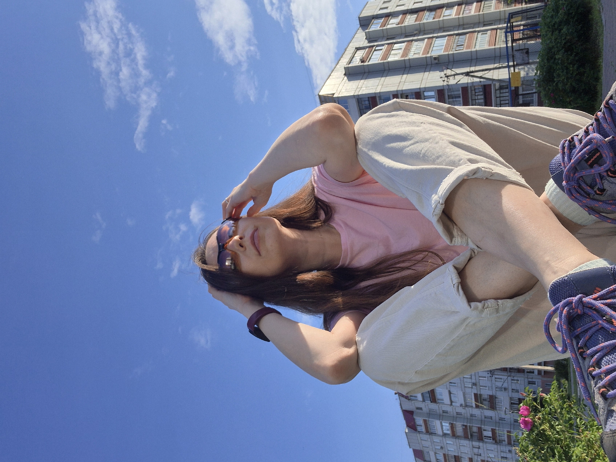 A low-angle, outdoor, medium shot captures a young woman sitting with her legs crossed, facing towards the right. She has long, brown hair and is wearing sunglasses, a pink t-shirt, and light-colored pants. Her right hand is resting on her head, while her left arm is bent, with her elbow resting on her left knee. A dark-colored smartwatch is visible on her left wrist. In the background, a tall, multi-story apartment building with many windows stands under a clear blue sky with scattered white clouds. To the left of the building, a dense green bush with pink flowers is visible. The foreground shows her legs and feet, with a pair of purple and orange laced sneakers.
