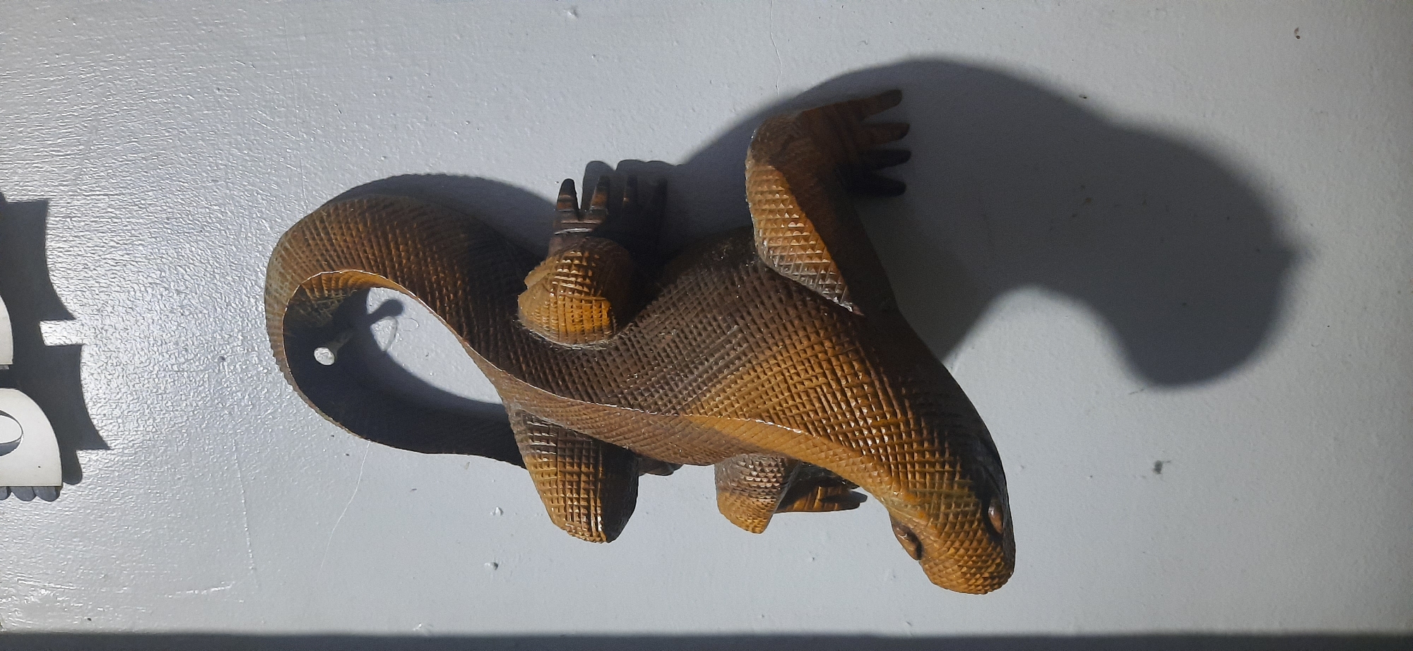 A carved wooden lizard hangs on a textured white wall in Cisauk, Indonesia. The lizard is oriented with its head to the left and its body curved upwards and to the right. Its scales are intricately carved with a diamond pattern, and its limbs are positioned as if it is clinging to the surface. A shadow of the lizard is cast on the wall to its right. In the upper portion of the image, a partial view of a white object with a dark shadow suggests other items on the wall or nearby. The lighting appears to be coming from the left, creating a distinct shadow.