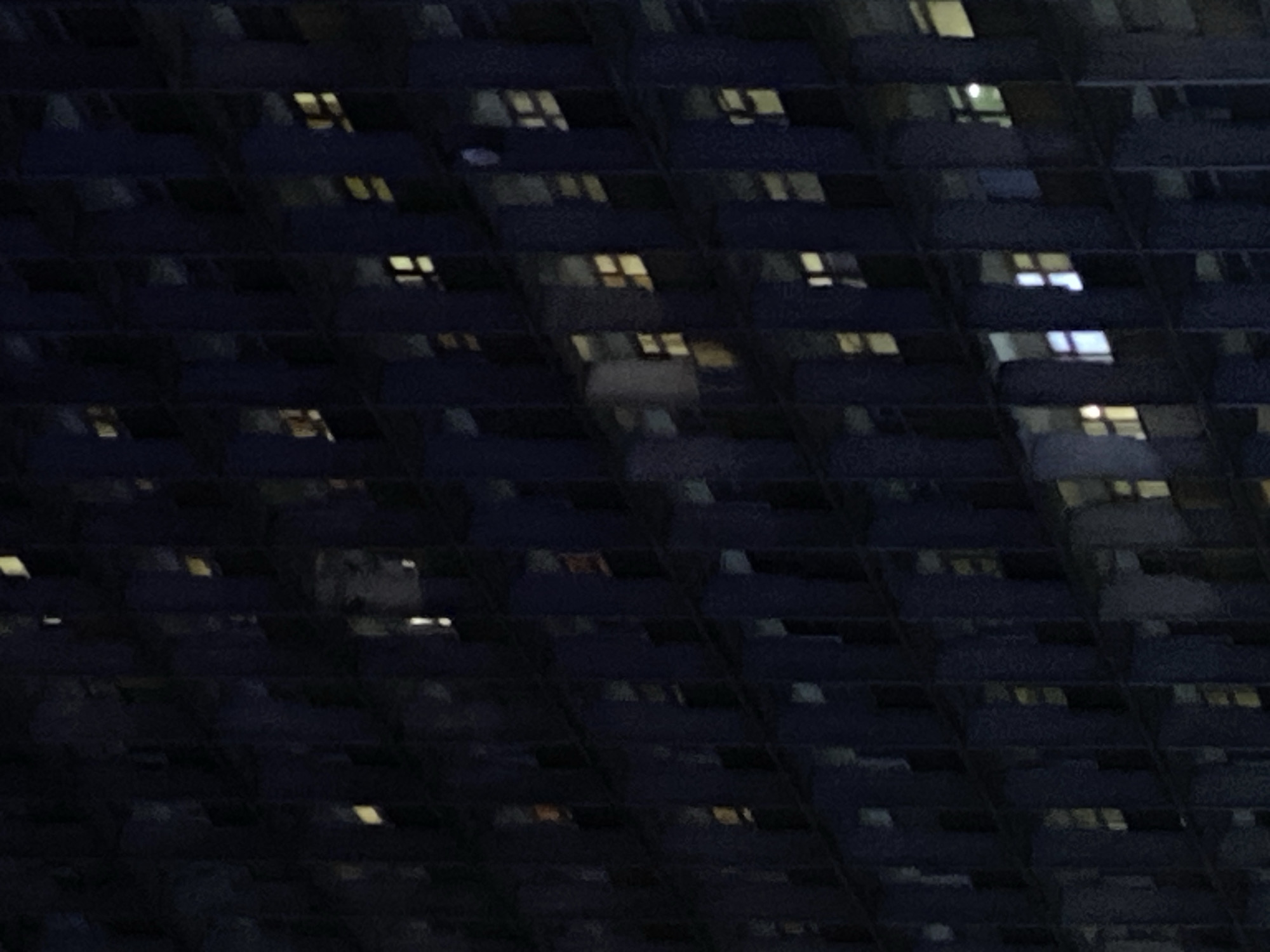 The image shows the exterior of a dark-colored building at night in Moscow, Russia. Light is visible in a number of windows, suggesting that the building is occupied. The facade appears to have a repeating pattern of horizontal elements.