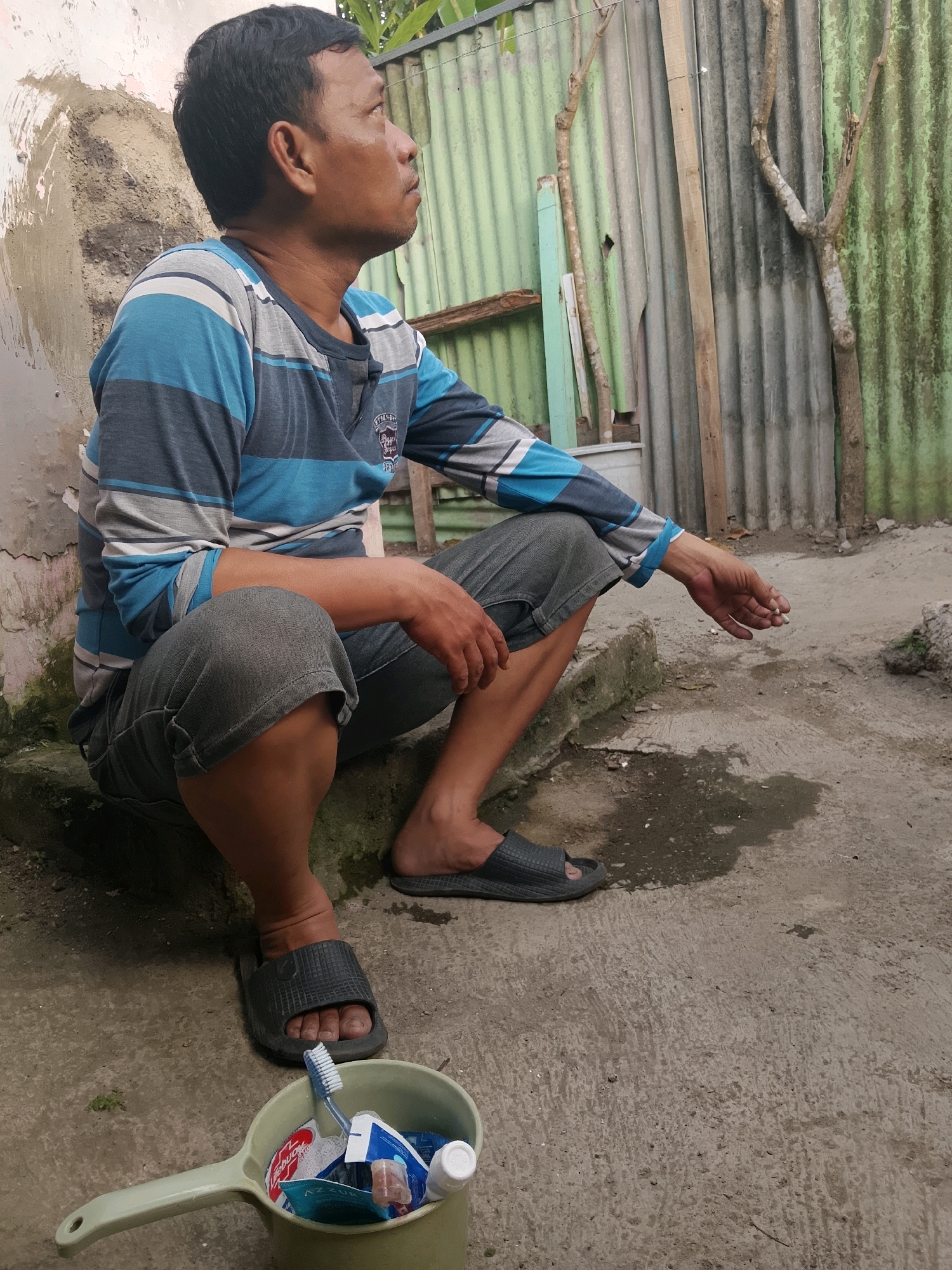 A man with dark hair and medium skin tone is squatting outdoors on a concrete surface next to a textured wall. He is wearing a long-sleeved shirt with horizontal stripes of blue, gray, and white, and dark gray shorts. His left hand is resting on his knee, and his right arm is extended with his hand near his mouth, holding a lit cigarette. In the foreground, a light green plastic scoop-like container is filled with toiletries, including a toothbrush, toothpaste tubes, and small packets. The background shows a corrugated metal fence, some wooden planks, and dry tree branches. The scene appears to be in an outdoor residential area. The specific location in Jember, Indonesia, is not discernible from the image alone.