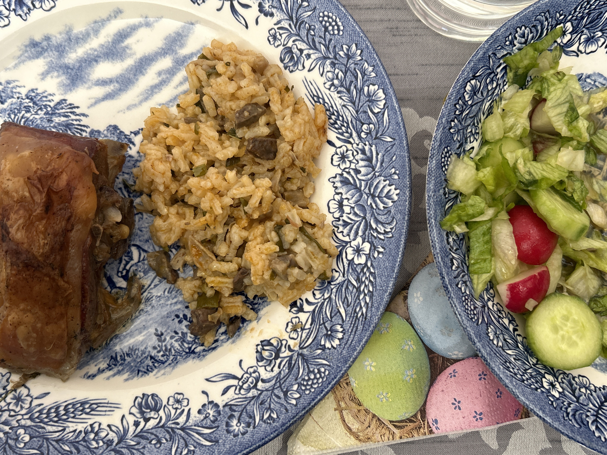 A close-up captures two blue and white floral patterned plates, one holding a portion of cooked meat alongside rice with chopped mushrooms or vegetables, and the other containing a fresh salad of lettuce, radishes, and cucumber. The plates rest on a surface with a design of painted easter eggs in the colors blue, green, and pink. The photograph appears to have been taken in an indoor setting, although the specific location in Krumovo, Bulgaria, is not discernible from the visual information provided.