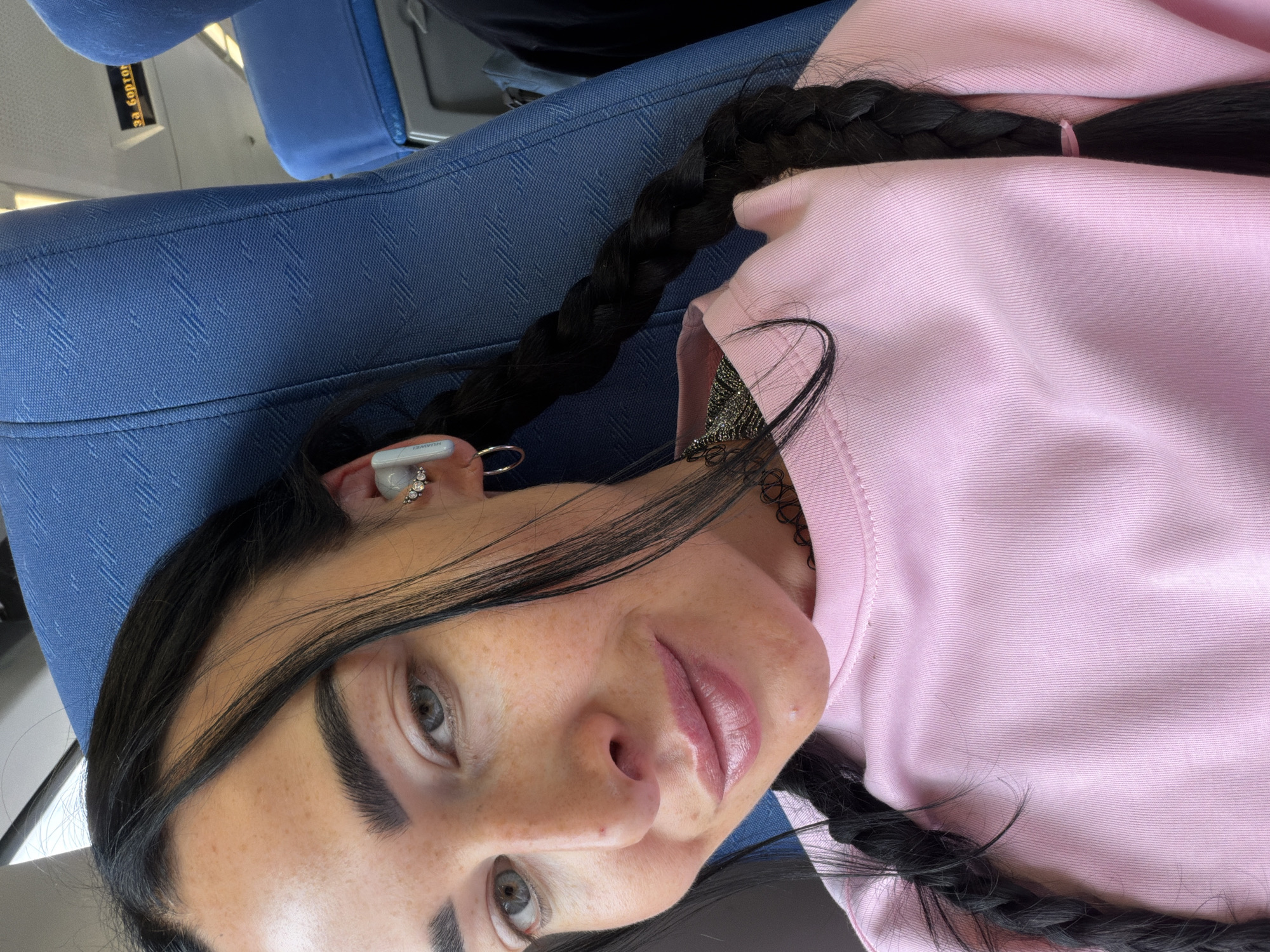 The media file is a close-up, selfie-style portrait of a young woman with dark hair styled in two braids, wearing a pink long-sleeved top. She is seated, and the background suggests an indoor setting, possibly public transportation given the blue upholstered seating visible. The lighting appears to be natural, indicating daytime. The woman has light-colored eyes, noticeable freckles across her face, and she is wearing small hoop earrings in her left ear. Her lips are glossy.
A small piece of white text is visible on what appears to be an earbud in her left ear, which reads "HUAWEI". There is also visible text on a yellow sign in the upper left corner of the image, in Russian: "ЗА БОРТОМ" (ZA BORTOM), meaning "Outside".
The overall mood is casual and relaxed, with the woman looking directly at the camera. The scene is likely within Russia, given the provided location context.