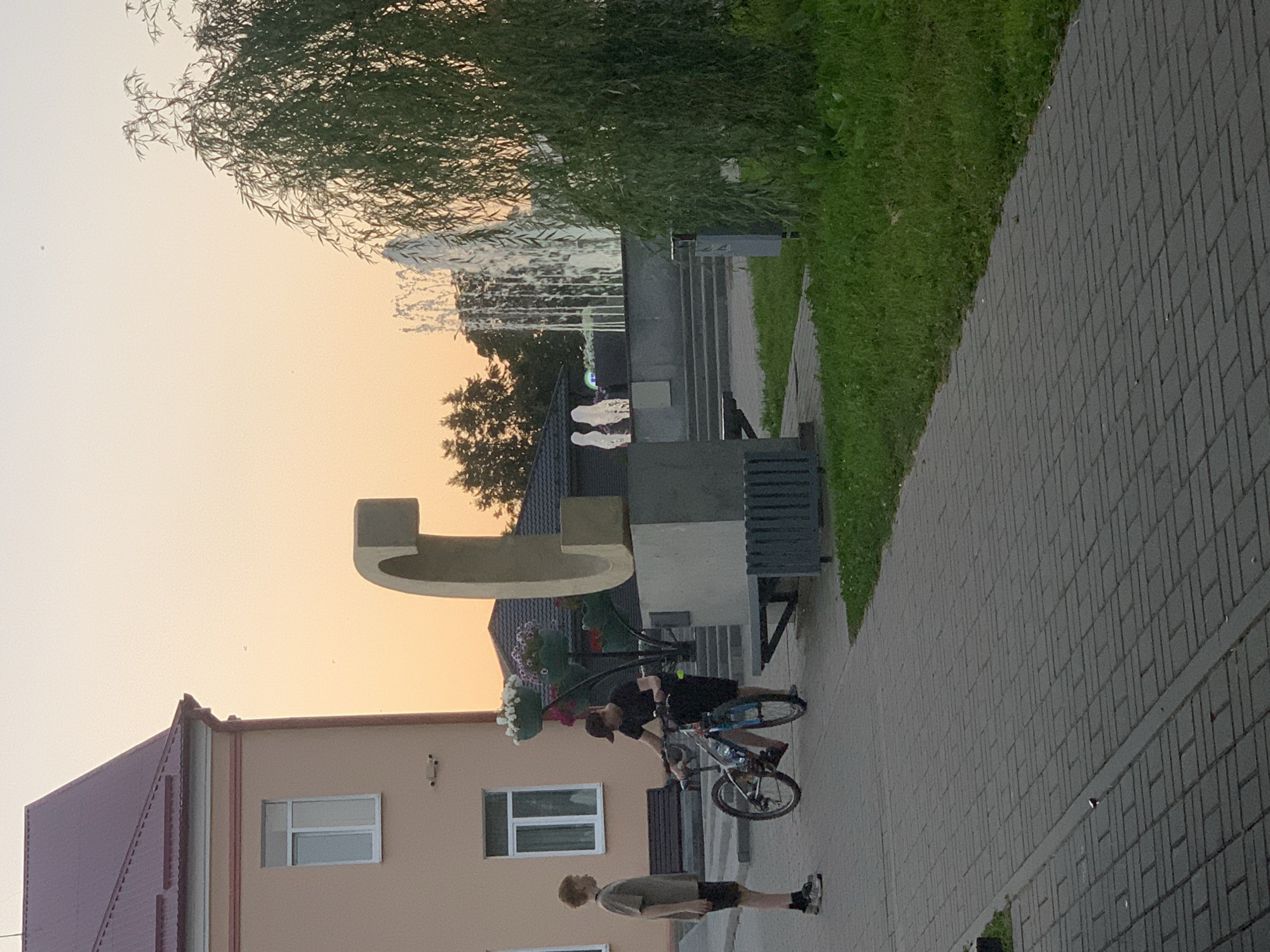 The image shows an outdoor scene in the city of Kapyĺ, Belarus, during what appears to be sunset or early evening. In the foreground, a paved walkway is visible. To the left, a light-colored building with a maroon roof and a window is present. Two young males are standing on the walkway. One, on the left, is wearing a gray t-shirt and black shorts. The other, to the right, is wearing a black t-shirt, black shorts, and a baseball cap, and is interacting with a bicycle. A large, curved, light-colored sculpture is a prominent feature in the mid-ground. Behind the sculpture, there are steps leading up to a plaza with a fountain spraying water. A weeping willow tree with dense foliage is on the right side of the frame. The sky is a pale orange, indicating the time of day.