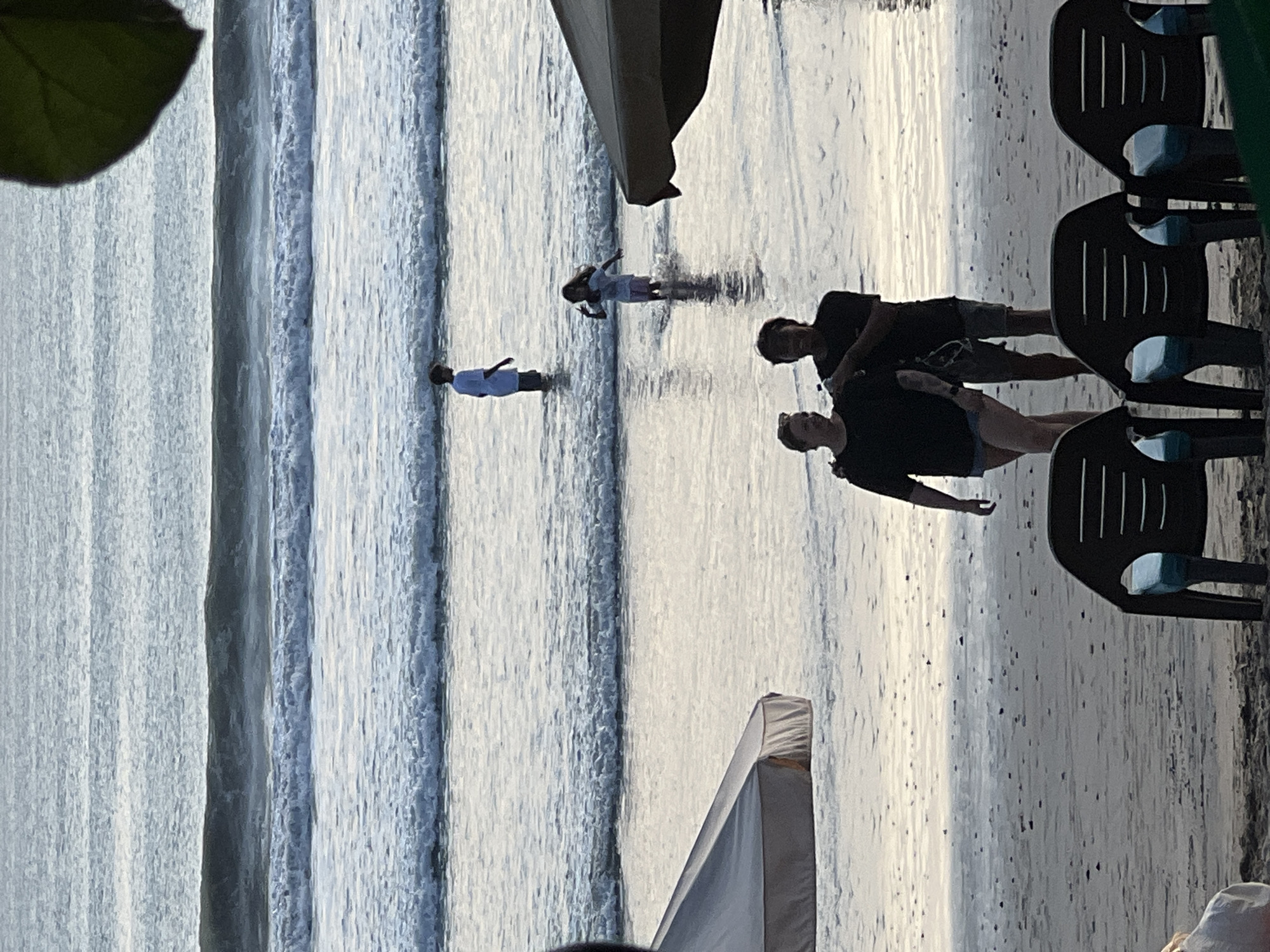 The image captures a serene beach scene in Kuta, Indonesia, during what appears to be either sunrise or sunset, given the soft, diffused lighting and the tranquil ocean waves. In the foreground, a couple walks hand-in-hand along the wet sand, their figures silhouetted against the gentle sea. Behind them, two children play in the shallow water, one a boy in a white shirt and shorts, the other a girl in a light-colored top and shorts, both seemingly enjoying the refreshing ocean. Further in the distance, more waves roll in, creating a rhythmic pattern on the water. Beach umbrellas are visible on either side of the frame, providing shade and adding to the relaxed atmosphere of a typical beach day. In the immediate foreground, empty beach chairs are set up, hinting at a leisurely day spent by the sea. The overall impression is one of peace and simple enjoyment in a beautiful coastal setting.