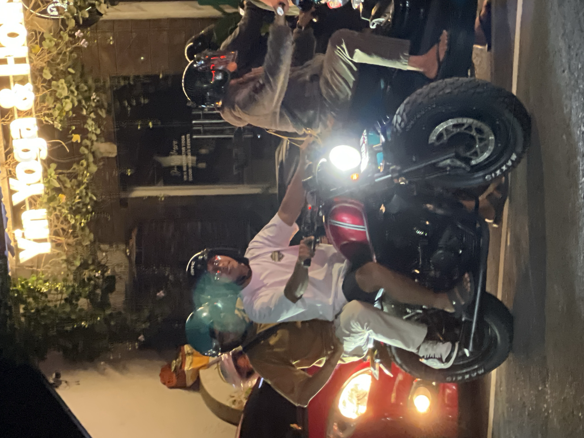 This nighttime photograph, taken in Ubud District, Indonesia, captures a street scene featuring three individuals on two motorcycles. The image is vertically oriented, showcasing the subjects and their surroundings under artificial illumination.
In the foreground, a person wearing a white t-shirt, light-colored pants, and a black helmet with a red stripe sits on a red and black motorcycle. The motorcycle's bright headlight is prominent, casting light forward. Their right foot, wearing a white sneaker, is visible near the front wheel. Directly behind this motorcycle, another person in a brown t-shirt and a blue helmet is partially visible, also seemingly on a motorcycle.
Further back and to the left, a third individual, dressed in a beige or light brown long-sleeved top and a black helmet, is seated on a dark-colored motorcycle, which also has a brightly lit headlight. The tire of one of the motorcycles is visible, clearly marked with the brand name "MAXXIS" on its sidewall.
The background reveals a building adorned with green foliage and a bright neon sign, partially displaying the text "Yin Yoga & Ho". The dark surroundings and prominent artificial lights indicate that the scene unfolds during the evening or night. The individuals appear to be either paused or in slow transit on the street.