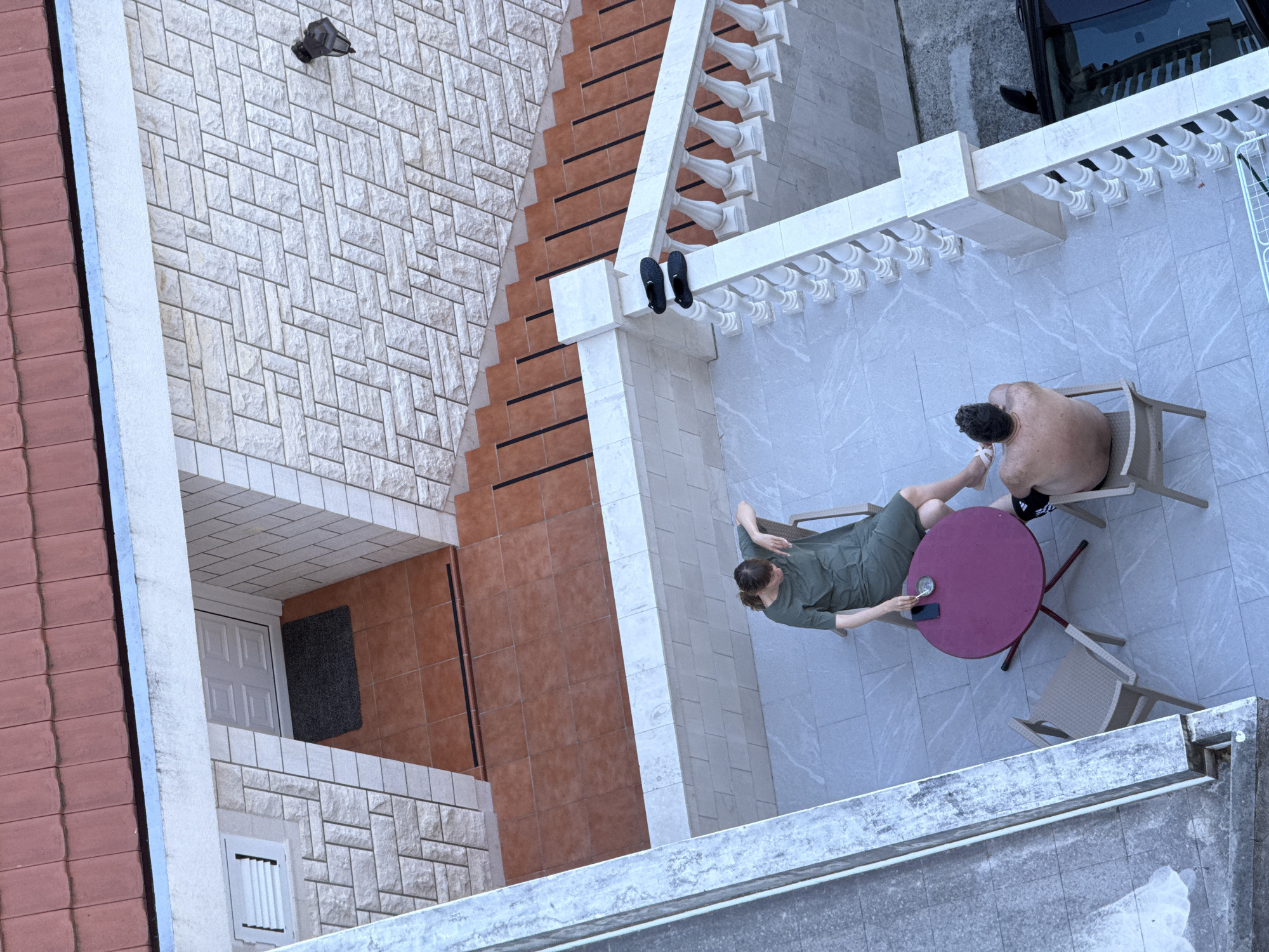 This is an aerial, medium-shot, outdoor, daytime photograph taken in Boreti, Montenegro. The image depicts a balcony area with two people, a man and a woman, seated at a round, magenta table. The woman, wearing a green dress, is holding a phone and an ashtray. The man, shirtless and wearing black shorts with a white stripe, is facing away from the camera.
The balcony is paved with light-colored, marbled tiles and features a white balustrade. To the right of the balcony, a black car is partially visible, parked next to a set of stairs. The stairs are made of reddish-brown tiles with black inlays and are bordered by white stone. Above the stairs and to the left, a wall is covered in light-colored stone blocks. A red-tiled roof is visible at the top of the frame. To the far left, a portion of another building's concrete exterior is visible.