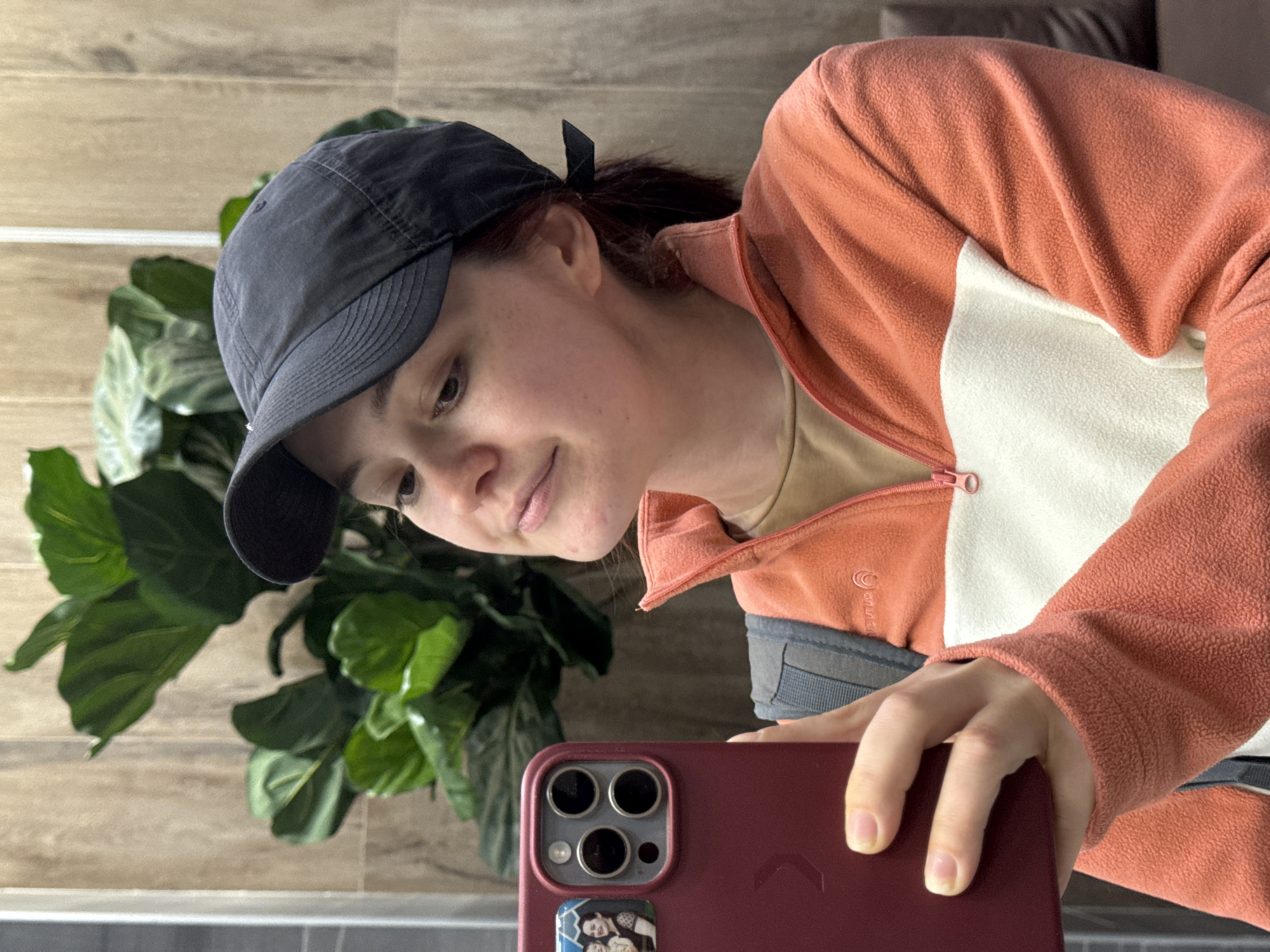 A person is taking a selfie indoors. They are wearing a dark blue baseball cap, an orange and white fleece jacket, and a beige t-shirt. The person is holding a red cell phone. There is a plant with large green leaves to the left of the person. The background includes a light-colored wood-paneled wall and a section of brown couch. The city of Izhevsk, Russia, is not discernible from the image.