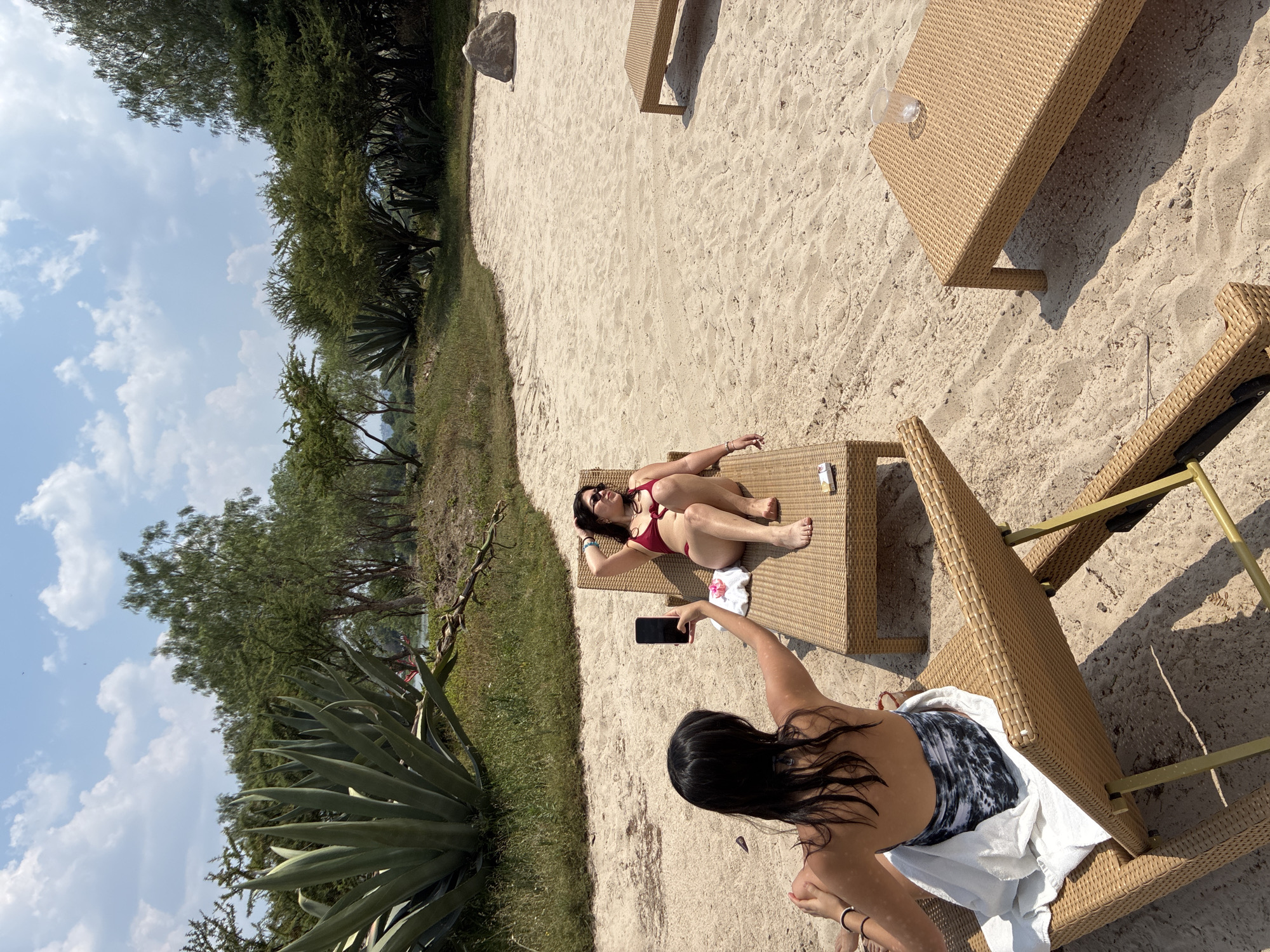 A woman in a bikini is lying on a lounge chair on a sandy beach in Pueblo Nuevo de Jasso, Mexico. Another woman stands near the lounge chair and holds a smartphone up to take a picture of her. There are other lounge chairs and a side table nearby. Trees and agave plants line the edge of the beach in the background. The sky is partly cloudy.