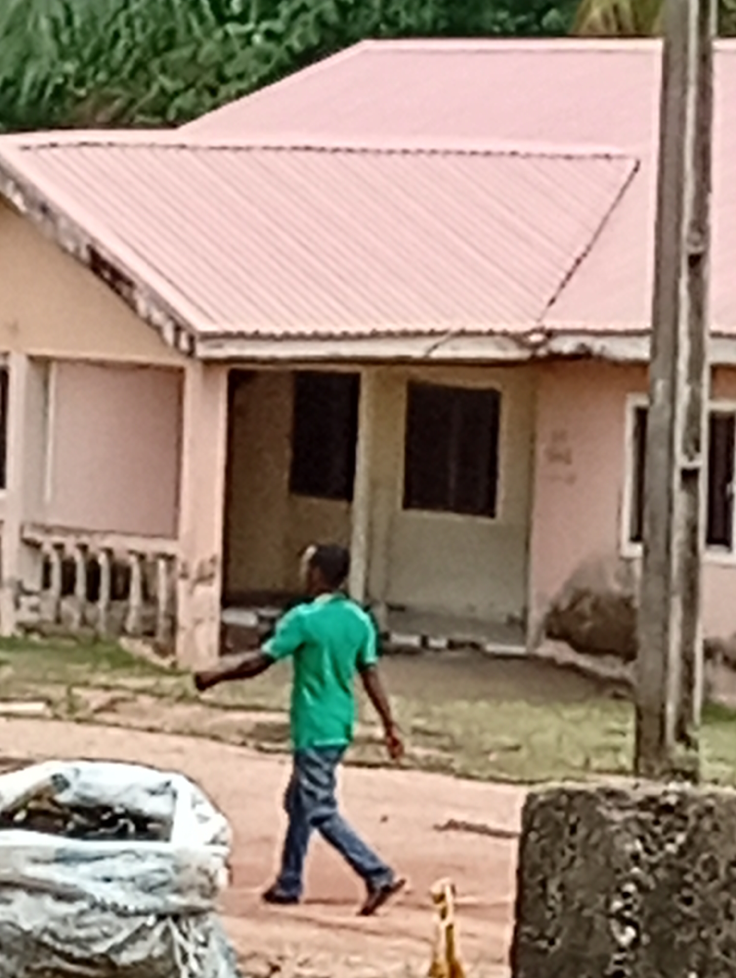 A man in a green shirt and blue jeans walks on a dirt road in front of a one-story building with a pink roof and a porch with railings in Unknown, Unknown. The building has two dark windows and an open doorway in the center. A utility pole is visible on the right side of the image, and a white bag of something sits on the left. A stone or concrete object is also present on the right. Trees are visible in the background.