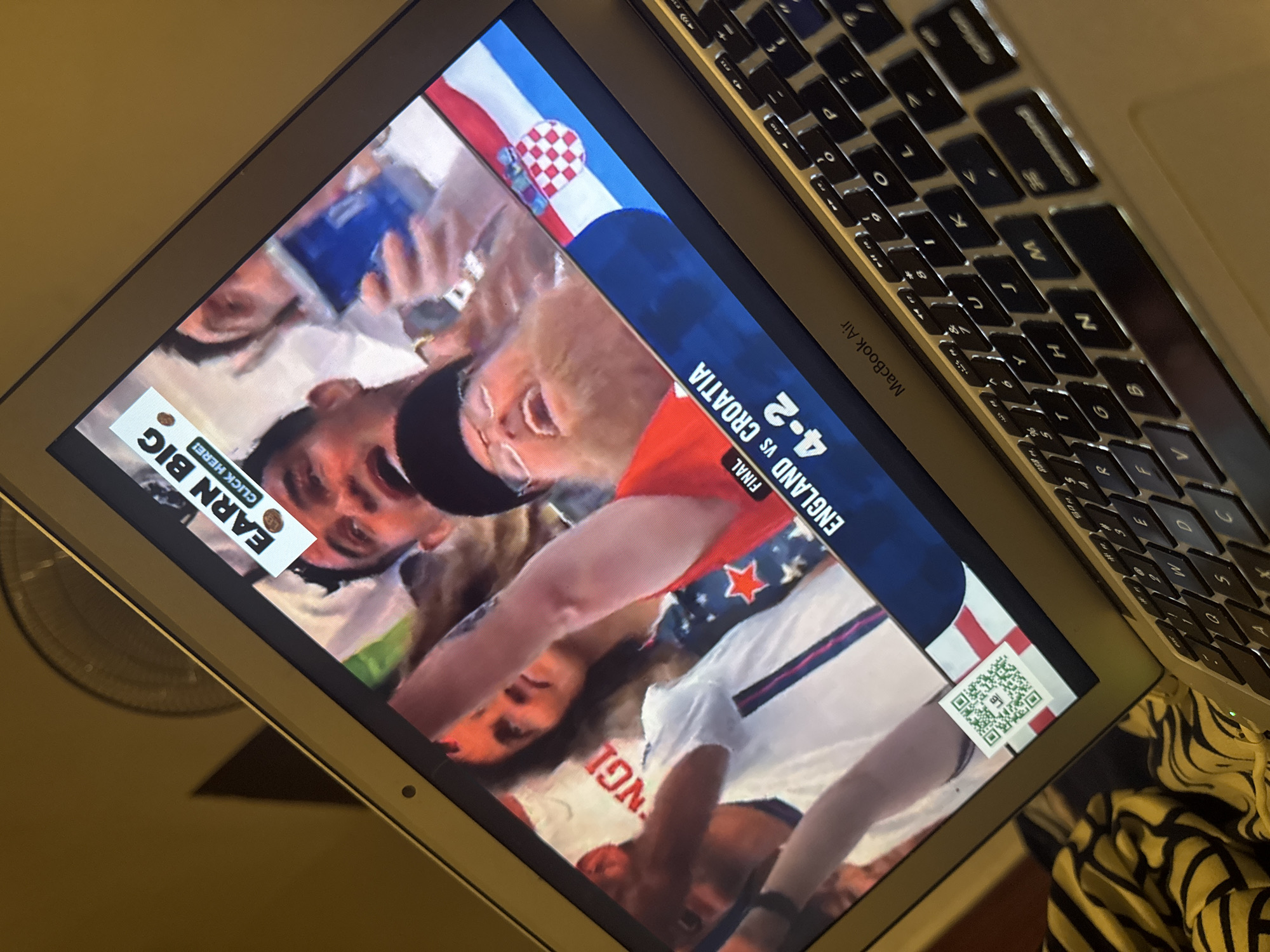 A laptop computer displays a sports match broadcast. The screen shows the score "ENGLAND VS CROATIA 4-2". Overlaying the broadcast is a white banner with "EARN BIG" and "CLICK HERE!". A QR code is also visible on the screen. The laptop is on a surface, with a portion of a black and yellow striped fabric in the lower left corner. The background is a plain, yellowish wall. The image is taken at a tilted angle, looking down at the laptop.