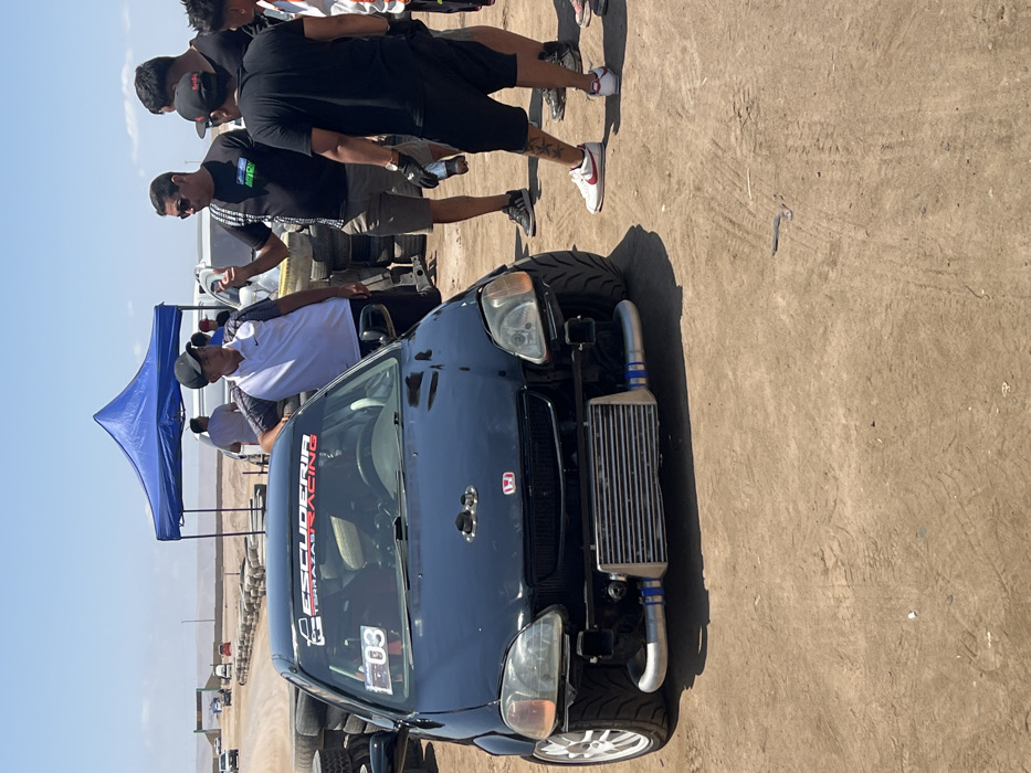 The image captures an outdoor scene under a bright, sunny sky, featuring a modified dark-colored Honda Civic surrounded by several men. The setting appears to be a race track, staging area, or car event, characterized by dry, sandy ground and distant barren hills or mountains under a clear sky.
In the foreground, a heavily modified dark blue or black Honda Civic hatchback is prominently displayed. Its front bumper has been removed or cut to expose a large, silver front-mounted intercooler with custom piping, indicative of a turbocharged engine setup. The car has wide, aggressive tires mounted on white multi-spoke alloy wheels. A red Honda badge is visible on the hood. Across the top of the windshield, a red banner reads "ESCUDERIA" in large white letters, with "TEAM AZAZ RACING" written below it. A white competition number plate with "03" in black is affixed below the banner on the passenger side of the windshield. The windshield itself appears somewhat dusty.
Several men are gathered around the car and in the background. To the left of the car, a man in a white short-sleeved polo shirt and a dark baseball cap is leaning into the driver's side window, possibly inspecting the interior or speaking with someone. Behind him and further left, a blue pop-up canopy provides shade, with another man partially visible underneath. To the right of the car, a group of men stand observing. One man in a black t-shirt and sunglasses is positioned near the car's front, looking down. His t-shirt has some white graphic elements. Another man, wearing a black t-shirt, black shorts, and white sneakers with red accents (possibly Nike Air Jordans), stands slightly behind him, revealing a tattoo on his lower leg. Other individuals are visible in the background, some in dark clothing, suggesting a team or group of spectators.
The overall atmosphere suggests an automotive event, such as a race, drift competition, or car meet, where performance vehicles are showcased and prepared. The bright sunlight casts strong shadows on the ground. Various pieces of equipment, possibly spare tires or tools, are visible on a cart or trailer in the mid-ground behind the group of men, further supporting the event context. In the far distance, beyond the blue canopy, some low barriers or structures can be seen, which might be part of a track or designated area.