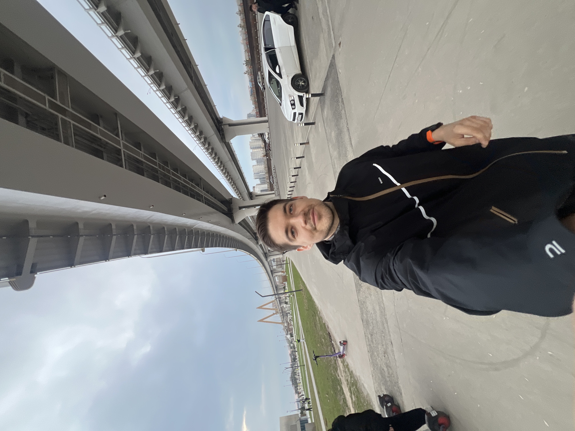 The image shows a man standing under a bridge in Kazan, Russia. He is wearing a black jacket and shorts, and he is looking at the camera. To his right is a white car. In the distance, there is a grassy area with scooters parked. The sky is cloudy.