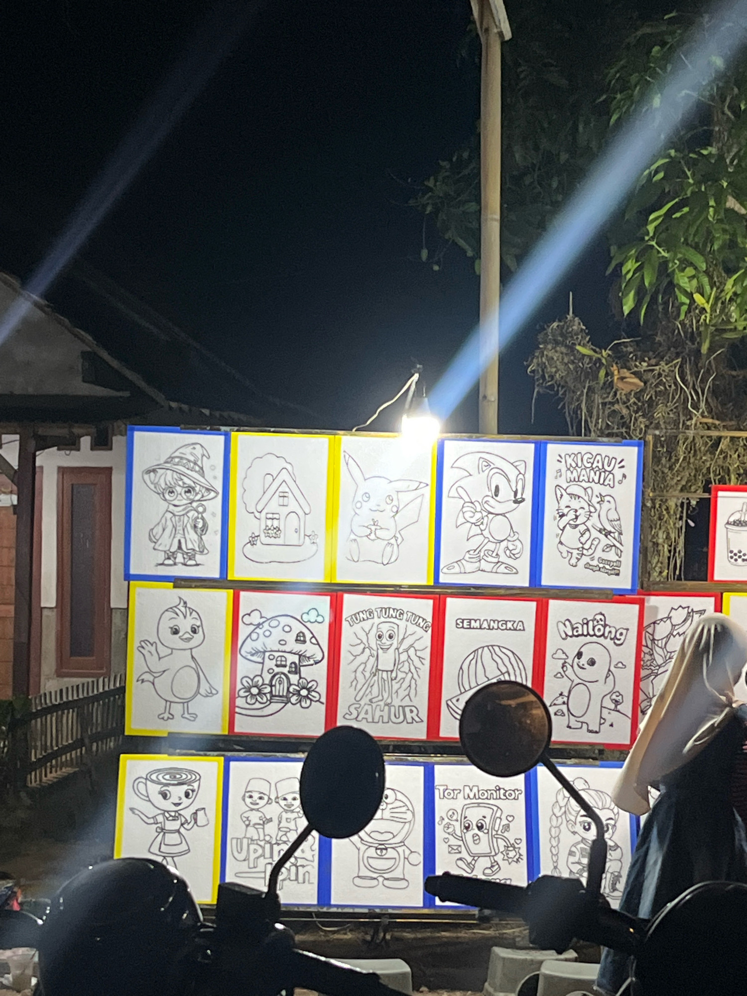 In the city of Sumberjo, Indonesia, at night, a display of children's coloring pages is set up outdoors. The coloring pages feature various cartoon characters, including a wizard-like figure, a house, Pikachu, Sonic the Hedgehog, a cat and bird, a chicken, a mushroom house, a watermelon, a baby-like character, a cup-shaped character, Doraemon, and a monitor character. The pages are arranged in rows on a wall, each framed by colored borders. A bright light shines down on the display, illuminating the area. In the foreground, the handlebars and mirrors of a motorcycle are visible, along with a person wearing a hijab. The background shows a building and trees.