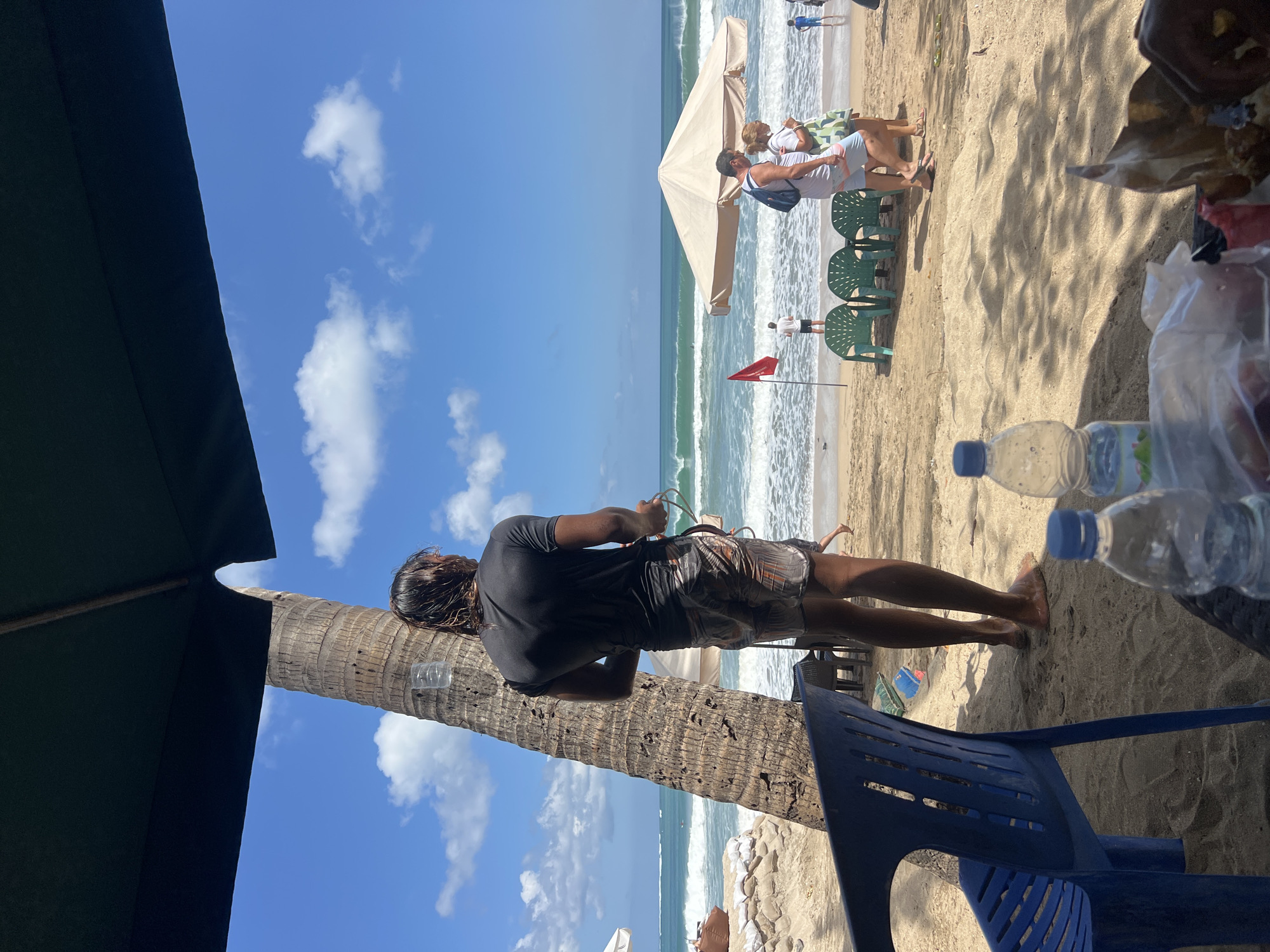 The image depicts a sunny day at Kuta Beach, Indonesia. In the foreground, a person stands near a palm tree, facing the ocean. They are wearing a dark t-shirt and patterned shorts, and holding a coiled rope. The beach has sandy ground with a few plastic chairs and a table with water bottles and food packaging visible. In the background, the ocean waves are breaking, and several people are seen walking along the shore, some under a large beach umbrella. A red flag is planted on the sand. The sky is blue with scattered white clouds.