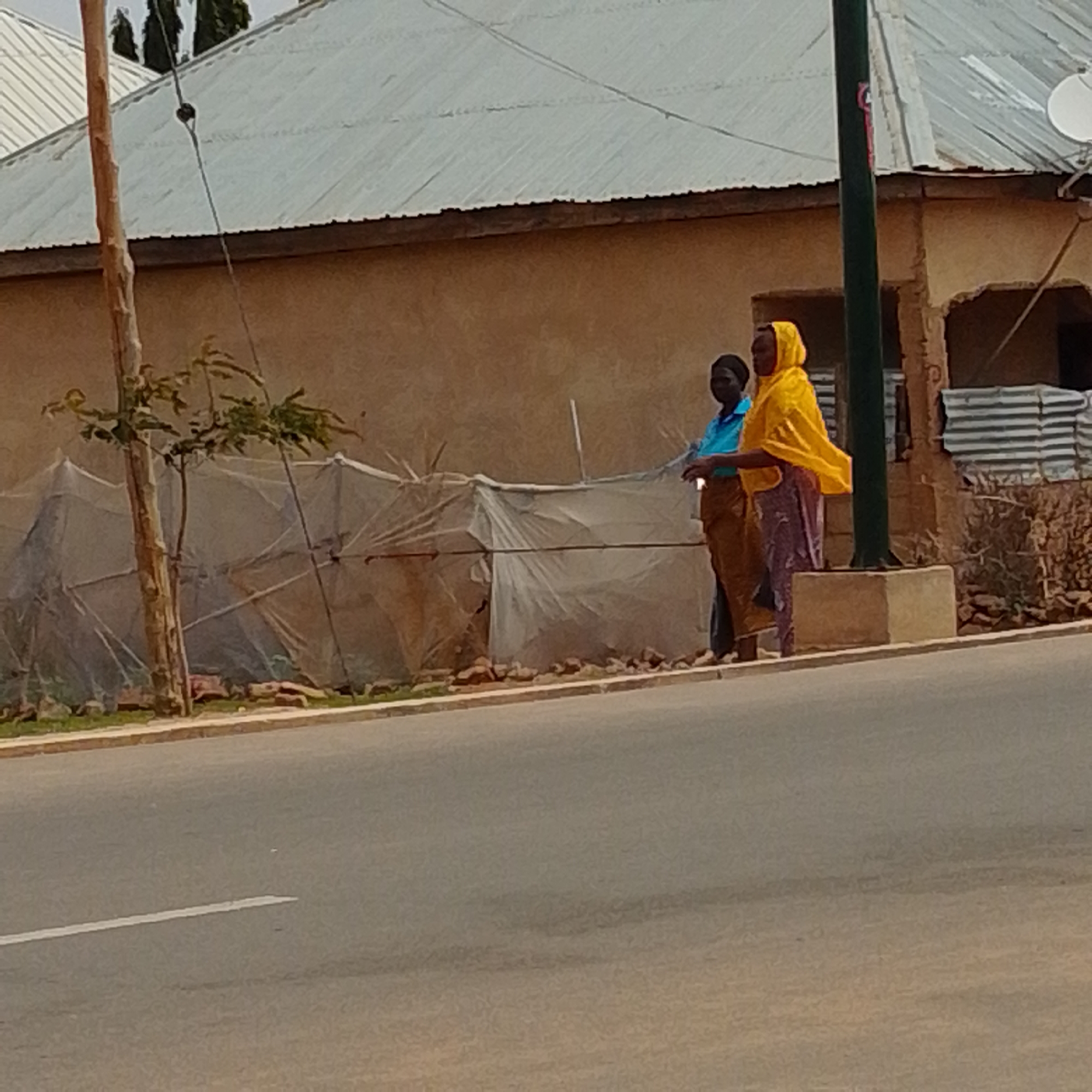 Two women are standing on a sidewalk next to a street. The woman on the left is wearing a blue shirt and a brown skirt. The woman on the right is wearing a yellow headscarf and a patterned skirt. Behind them is a building with a corrugated metal roof and a stucco wall. There is also a tree and some plastic sheeting. The street is paved and has a white line painted on it. The scene appears to be in a sunny outdoor environment.