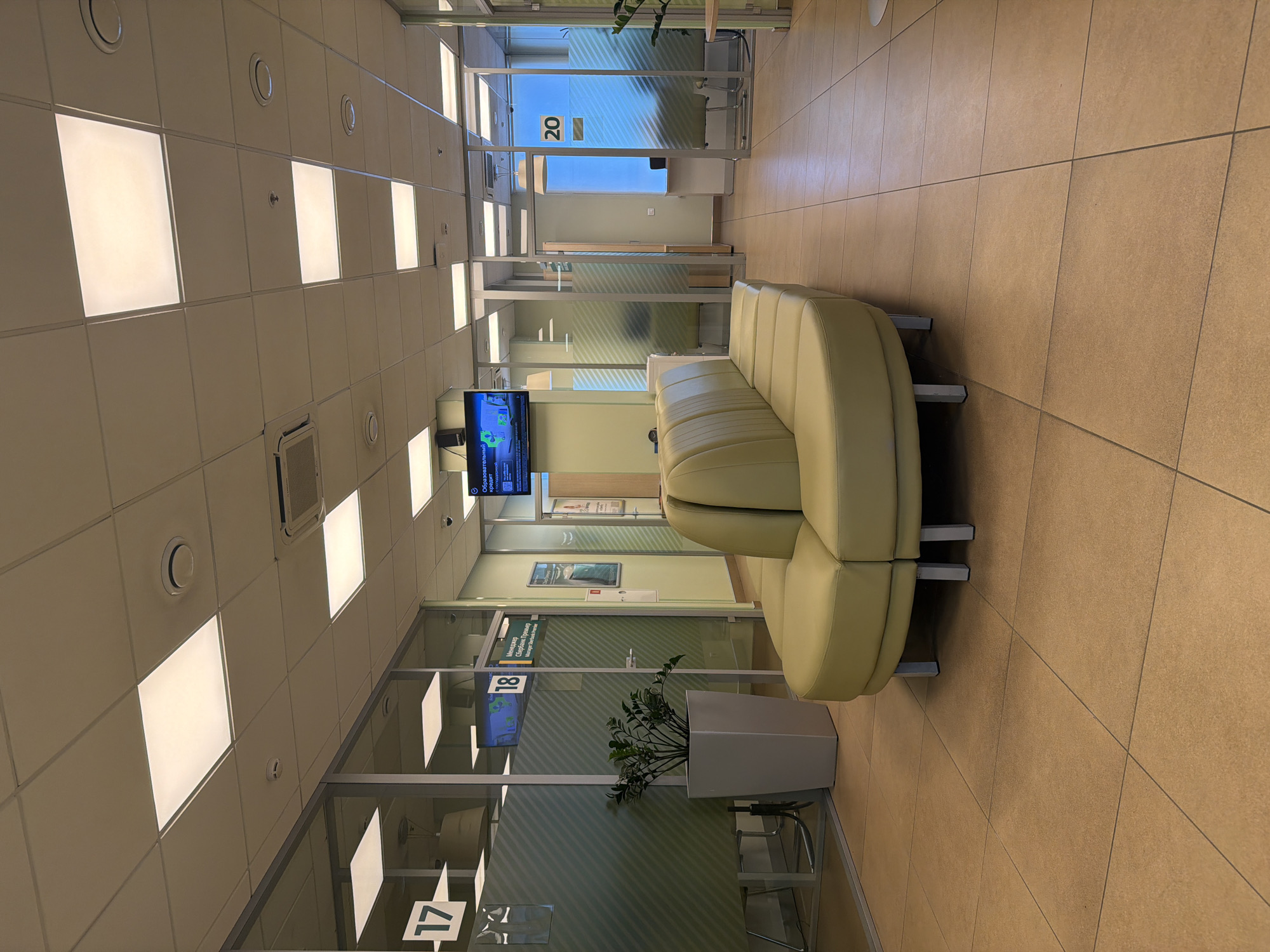 The image depicts the interior of a bank or service center, likely in Krasnoyarsk, Russia, based on the architectural style and interior design common in Russian commercial spaces. The scene shows a waiting area with a pale green, curved sofa placed in the center of a hallway. The floor is covered in beige tiles. The walls are mostly glass partitions, indicating separate offices or service windows. Signage with numbers (17, 18, 20) is visible on the glass walls, presumably to identify the different service points or offices. A television screen is mounted on the wall, displaying information or advertisements. A large, square planter containing a plant sits near the sofa. The ceiling is a drop ceiling with evenly spaced, square light fixtures. No people are visible in the immediate foreground. The overall environment suggests a clean, modern, and well-lit commercial space.