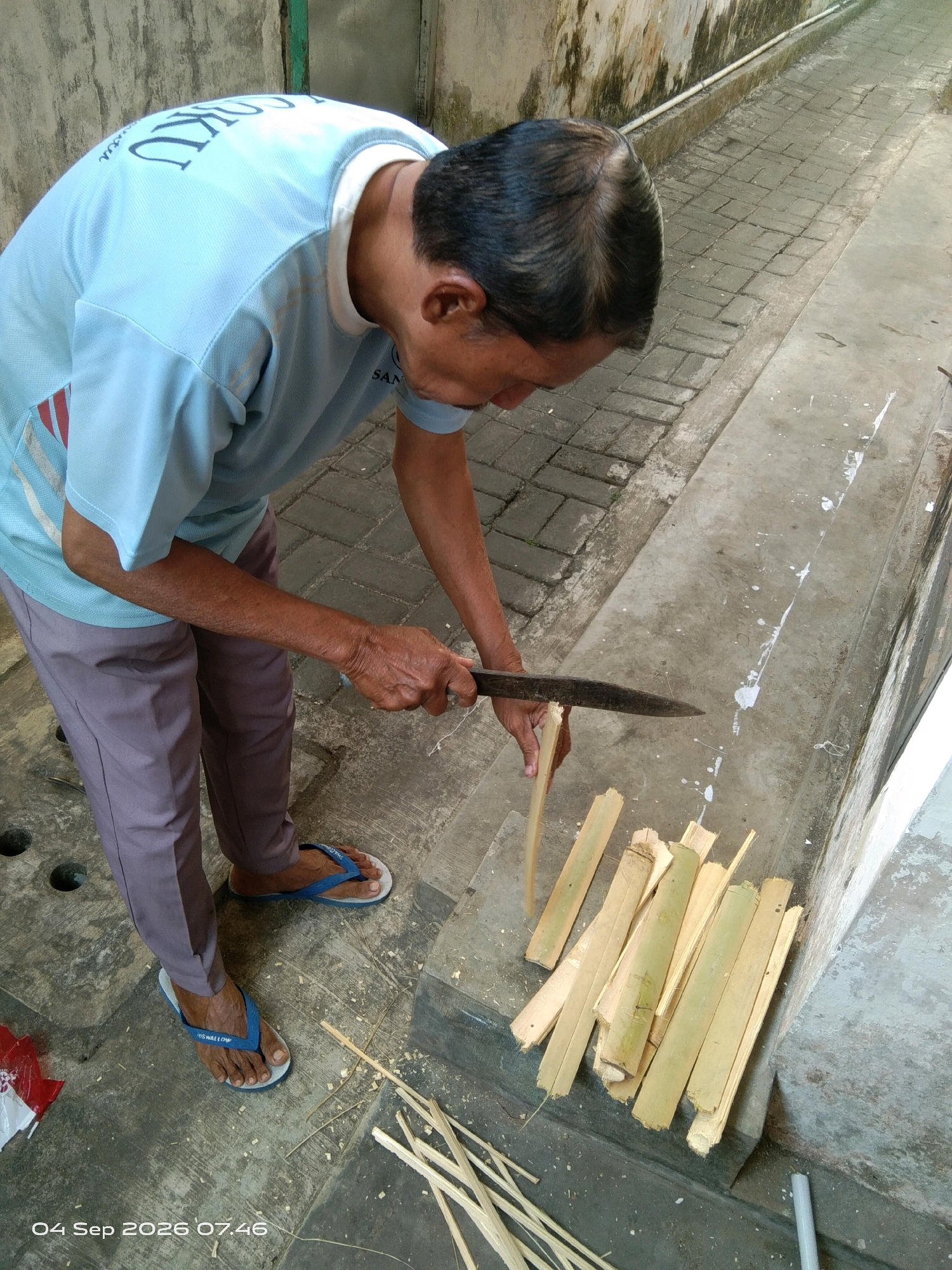 An Indonesian man is shown from the chest down, bent over and using a large knife to cut thin strips of bamboo. He is wearing a light blue t-shirt and grey pants, and blue flip-flops. The bamboo strips are piled to his right. The scene appears to be outdoors on a paved walkway. A wall is visible on the left, with a green pipe running vertically. A cobblestone path runs behind the man and to the right. The ground is concrete, with some white paint splatters. The image is dated "04 Sep 2026 07:46".