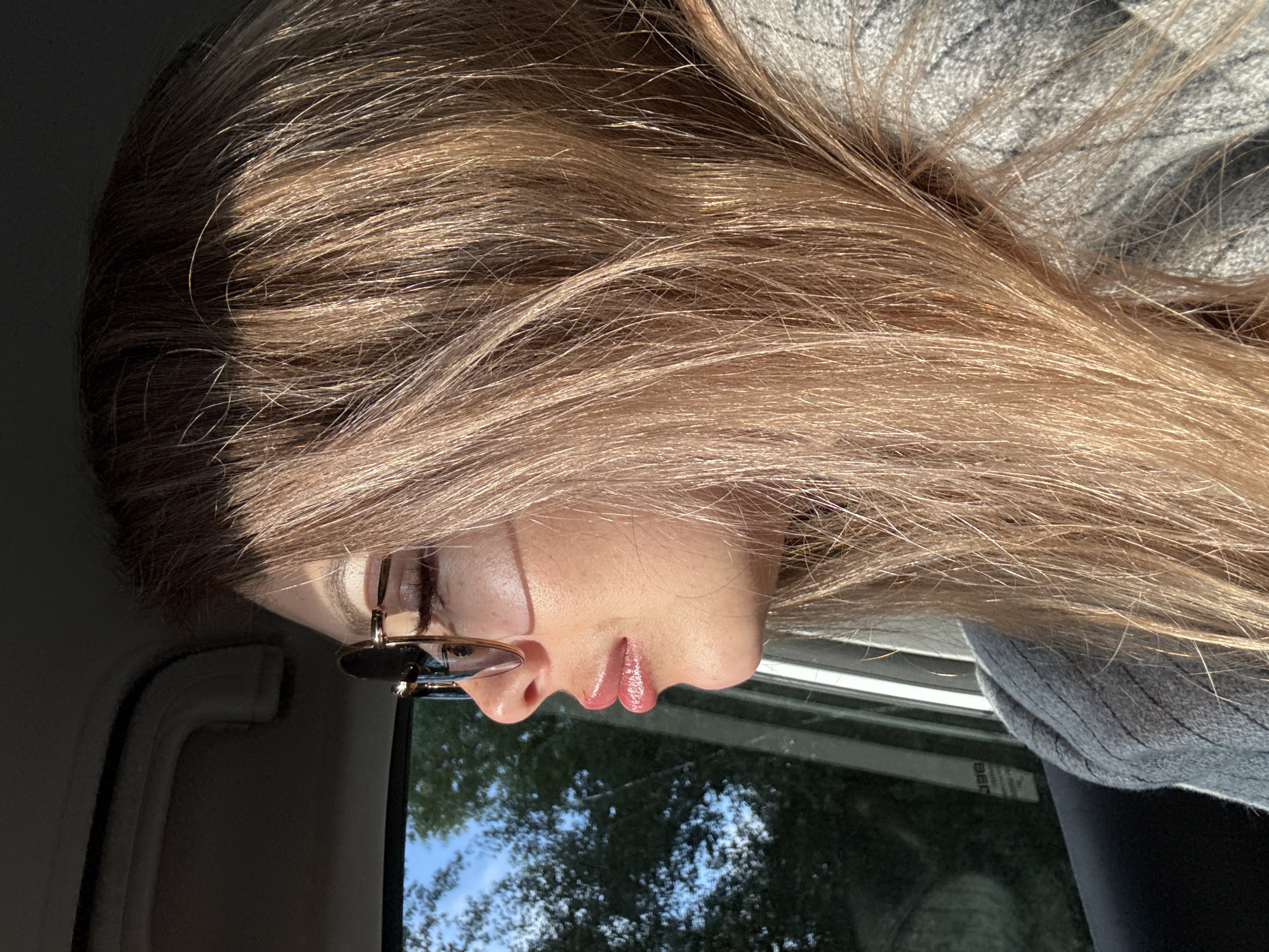 A woman with long, light brown hair and sunglasses is sitting in a car, looking out the window. The sun is shining, casting shadows on her face. She is wearing a grey sweater. The background shows trees and a blue sky. The location is unknown.