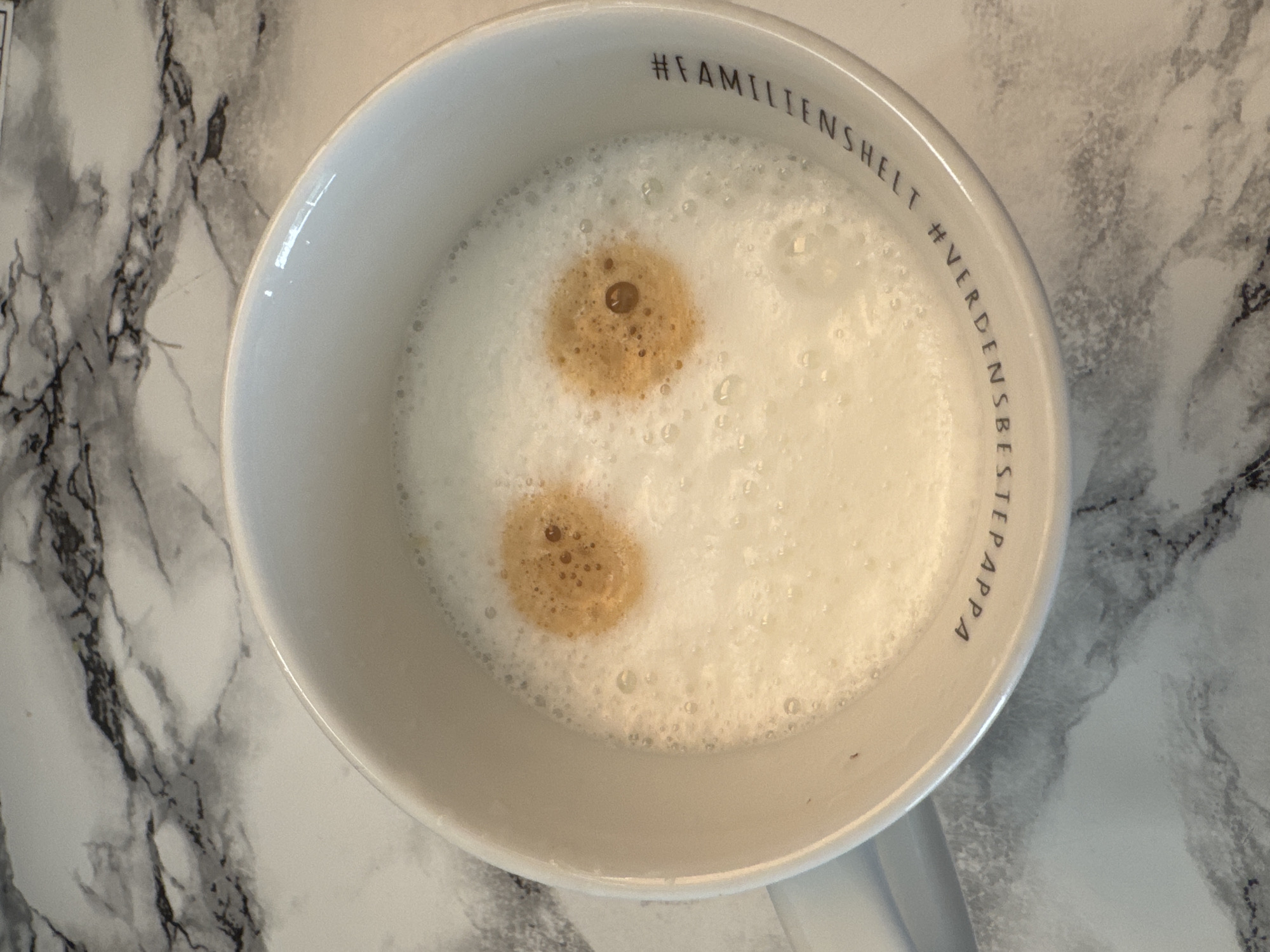 An overhead, medium shot shows a white mug filled with a frothy, light-colored beverage, likely a latte or cappuccino, resting on a white marble-patterned surface. Two circular dollops of darker liquid, resembling coffee or espresso, are positioned in the foam, forming what appears to be eyes. The mug has text printed in black around its rim. The text reads "#FAMILIESHELT #VERDENSBESTEPAPPA". The scene is indoors, possibly in a kitchen or dining area, in Moelv, Norway. No people are visible in the image.