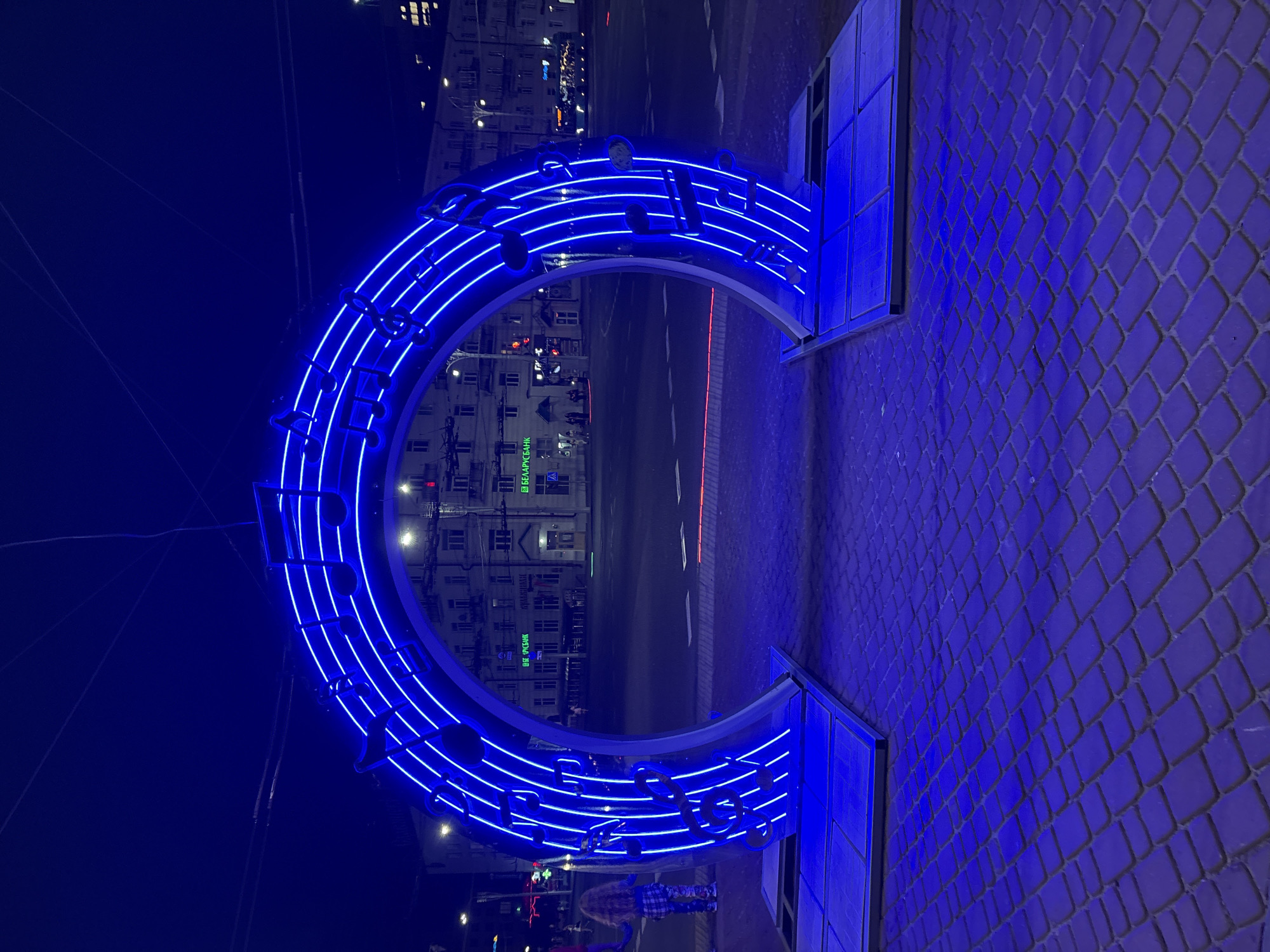 The image shows a large, blue neon archway decorated with musical notes and staves, positioned on a cobblestone sidewalk at night. The archway is located in front of a street with buildings in the background, some of which are illuminated. A person with long, blonde hair and wearing checkered clothing is standing to the left of the archway, partially obscured from view. The lighting suggests an urban environment, possibly a public space or a street in the city of Vitsebsk, Belarus.