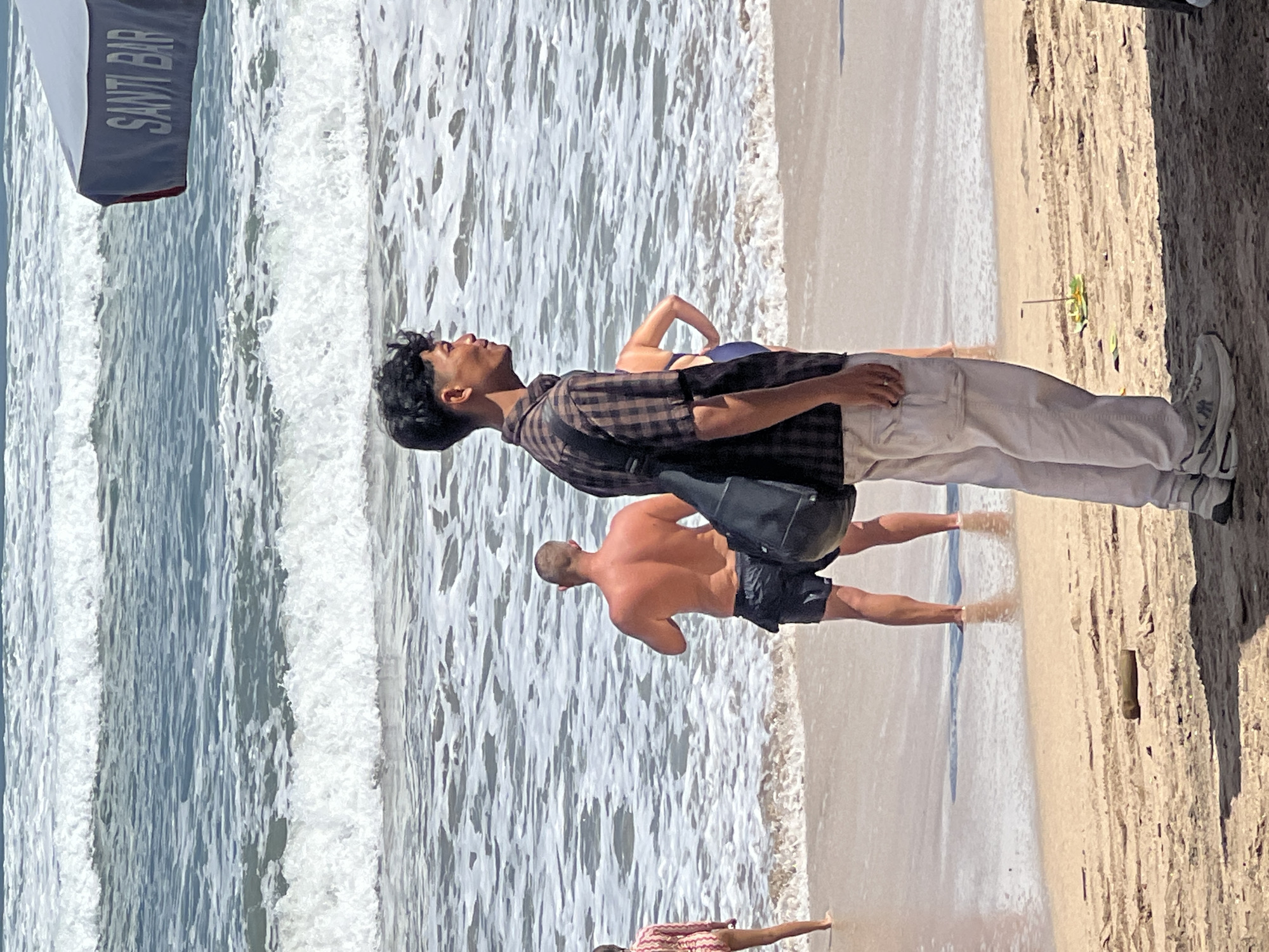 A young man in a plaid shirt and light-colored pants stands on a beach in Kuta, Indonesia, facing away from the camera and looking out at the ocean. He has a black bag slung over his shoulder. In the background, waves crash on the shore. A man in black swim trunks stands in the shallow water, and a woman in a blue swimsuit is partially visible behind the young man. Further in the background, a beach umbrella with "SANTI BAR" printed on it is visible. The sand is light brown and strewn with debris, including a piece of driftwood and a small, colorful object stuck in the sand.