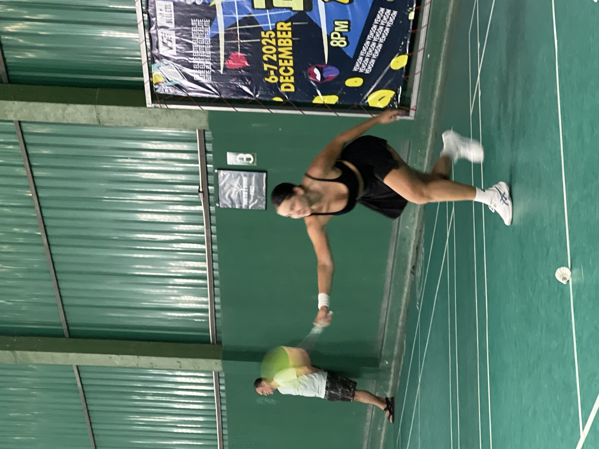 In a badminton court in Ban Bang Rak, Thailand, a young woman in a black sports bra and black skirt is captured in motion, playing badminton. She is in the process of hitting a shuttlecock with her racket, her body contorted in a dynamic pose. Her white athletic shoes are laced up, and white socks are visible. A blur of motion suggests the swiftness of her movement.
In the background, a man in a light-colored t-shirt and patterned shorts leans against the green wall of the court, observing the game. He is wearing dark sandals. The court itself is marked with white lines on a green surface.
To the right of the image, a large banner is affixed to a pole. The banner is predominantly black with yellow and red accents. It displays text including "6-7 2025 DECEMBER" and "8PM", as well as repeated text "VENSON VENSON VENSON VENSON". The wall behind the players is also green, with a corrugated texture. A metal pole is visible in the upper left portion of the image, part of the court's structure. A small sign with a "B" on it is mounted on the wall behind the male player. The ceiling structure is visible at the top, with green corrugated panels and metal beams.