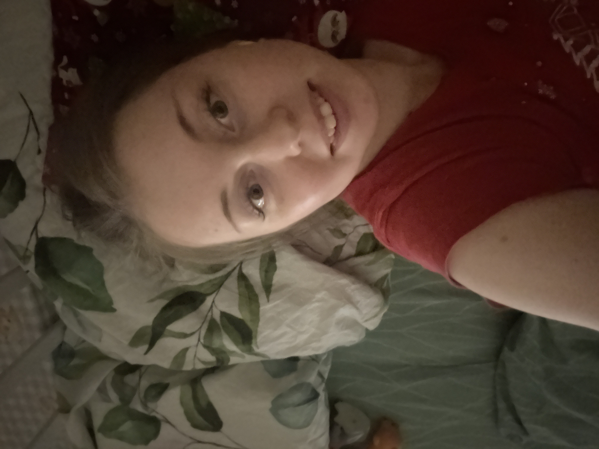 A person is lying on a bed with a leafy-patterned pillow. The person is wearing a red shirt and smiling. The location is unknown.