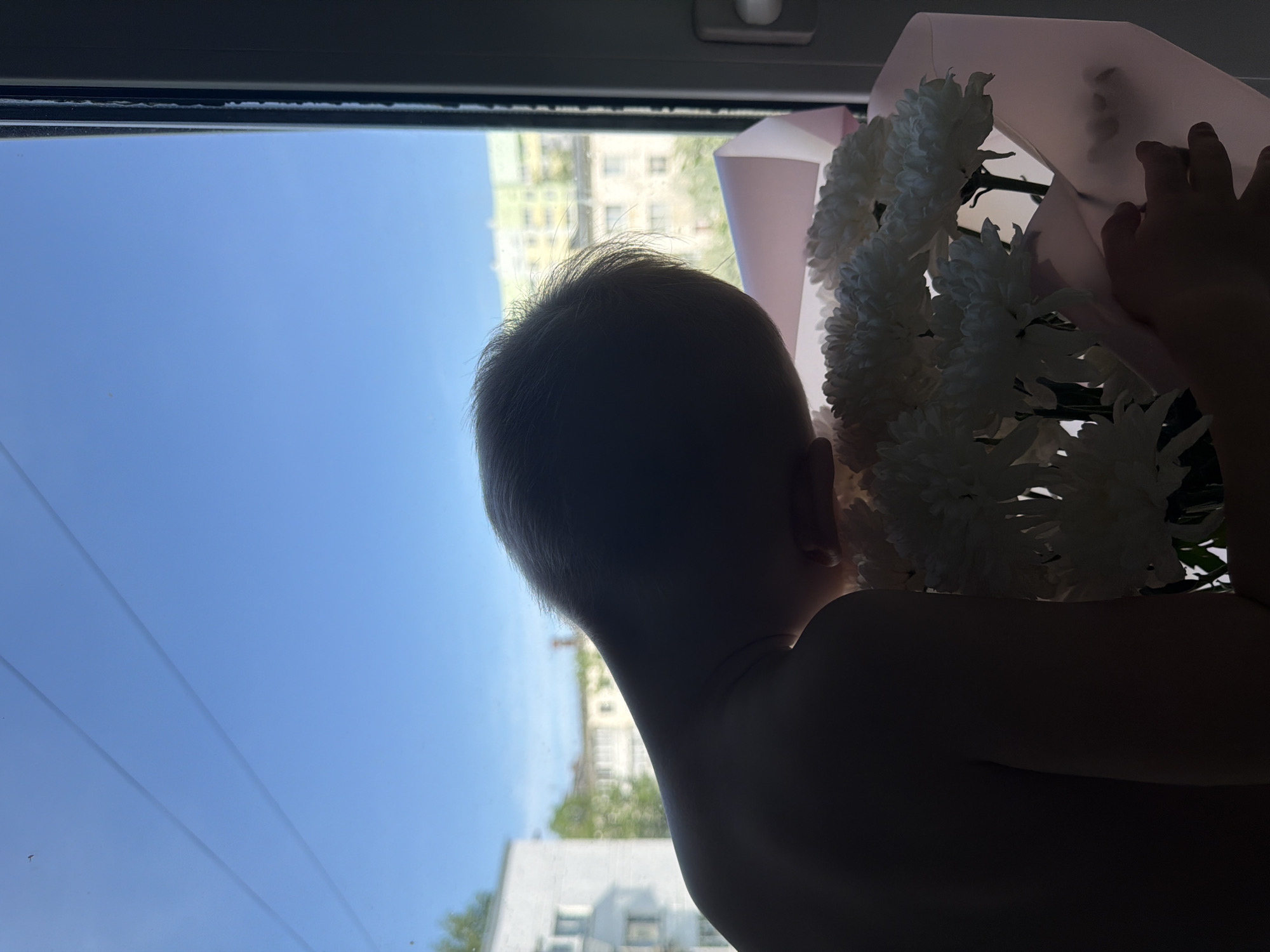A low-angle, indoor shot presents a child holding a bouquet of white chrysanthemums against a window view. The child is seen from the back and side, their body silhouetted. They are holding the flowers in a pink wrapping. Through the window is a blue sky and buildings, the location appears to be Chaikovsky, Russia.