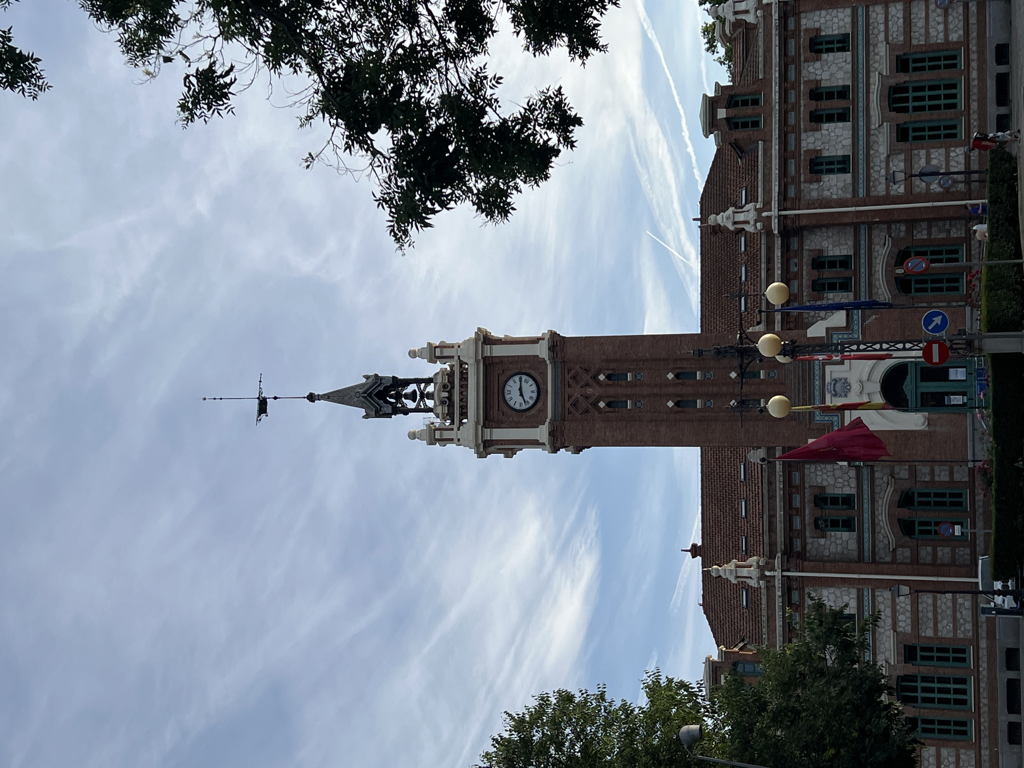 The image depicts the Ayuntamiento de Aranjuez, the city hall of Aranjuez, Spain, located near Madrid. The building is characterized by its red brick and stone facade with green window frames. A prominent clock tower stands tall, with a weather vane at the top. Several flags are displayed in front of the building. The sky is partially cloudy with contrails from airplanes. Tree branches frame the top and bottom of the image.
