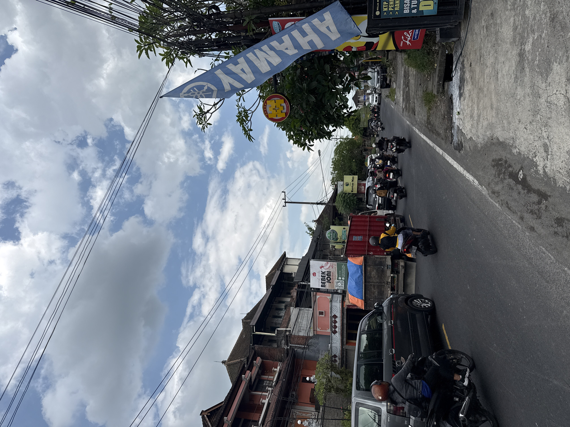 The image captures a street scene in Ubud District, Indonesia. A line of motorcycles and cars are traveling on the gray asphalt road. A blue banner with the word "AHAMAY" is visible. Buildings with signage like "BEBEK JONI" and "Kertas Mas" flank the road, displaying local businesses. Power lines crisscross above the scene. The sky is partly cloudy.