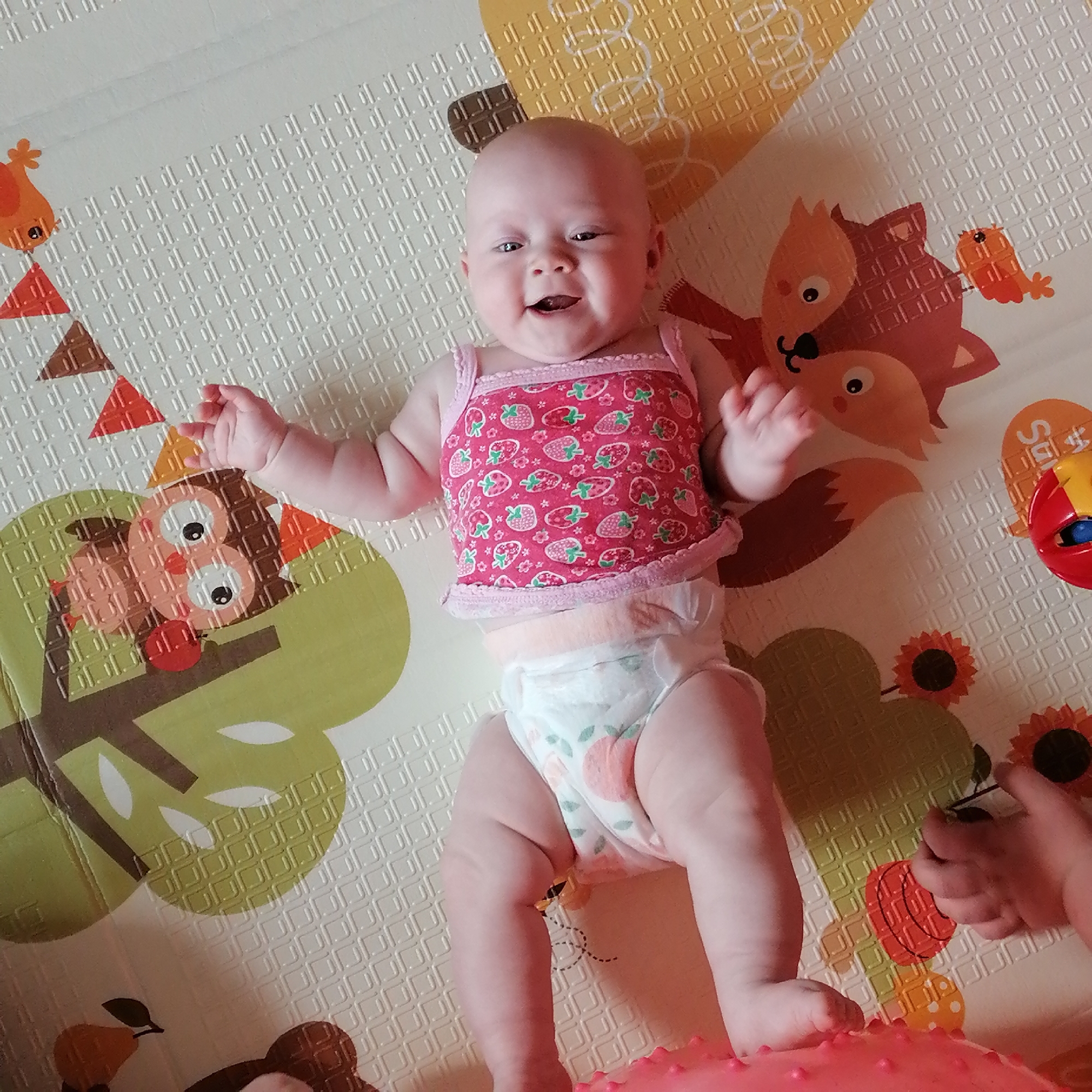 An overhead shot shows a baby lying on a playmat with a pattern of woodland creatures. The baby is wearing a pink strawberry-print tank top and a diaper. The baby is smiling and looking up at the camera. A child's hand is visible on the right side of the frame, reaching towards the baby. The playmat has a textured surface with embossed squares. The baby is positioned in the center of the frame, with the playmat extending outwards in all directions. The lighting is bright and even, indicating the photo was likely taken indoors or in a shaded outdoor area. The background is a continuation of the playmat's design. The overall impression is one of a cheerful and playful scene.