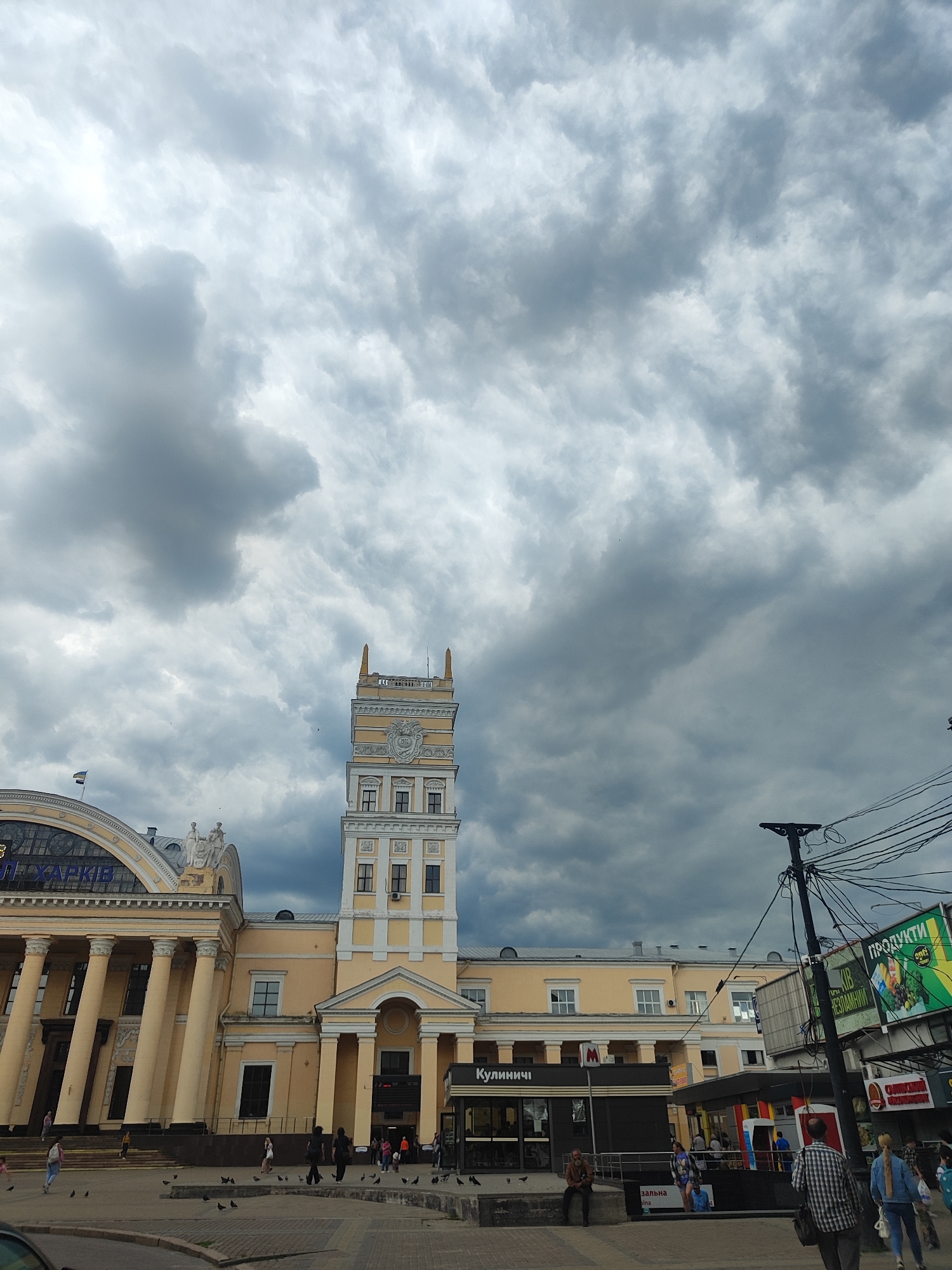 A low-angle, wide shot captures the facade of a large, pale yellow building with neoclassical architectural elements, likely a train station, under a heavily overcast sky. A prominent clock tower with decorative molding and a tiered roof rises from the center of the building. To the left, a curved section of the building features a row of tall, fluted columns. Several people are visible on the paved area in front of the building, some walking and some standing. Pigeons are scattered on the ground. A large, dark-gray cloud formation dominates the sky. In the right foreground, a dark utility pole with numerous wires is visible, along with a green billboard advertising "Продукти" (Products) and a smaller red and white sign. The ground is paved with gray concrete slabs.