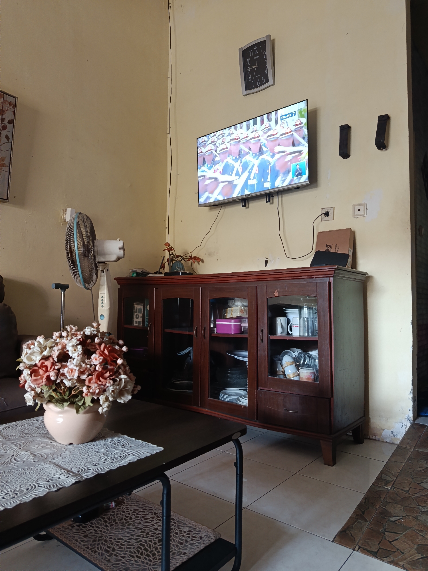 The image depicts an interior scene of a room, possibly a living room, in a home in Keboananom, Indonesia. The walls are a light yellow, and the floor is tiled. A dark wooden cabinet with glass doors stands against the wall, filled with various items including plates, cups, and what appear to be glassware. On top of the cabinet, a flat-screen television is mounted on the wall, displaying a broadcast showing a marching band in uniform. To the left of the television is a square wall clock showing the time. A standing fan is positioned to the left of the cabinet. In the foreground, a dark coffee table is set with a lace-patterned placemat and a vase filled with artificial flowers in shades of pink and cream. A dark-colored sofa is partially visible in the background. The overall impression is of a lived-in domestic space.