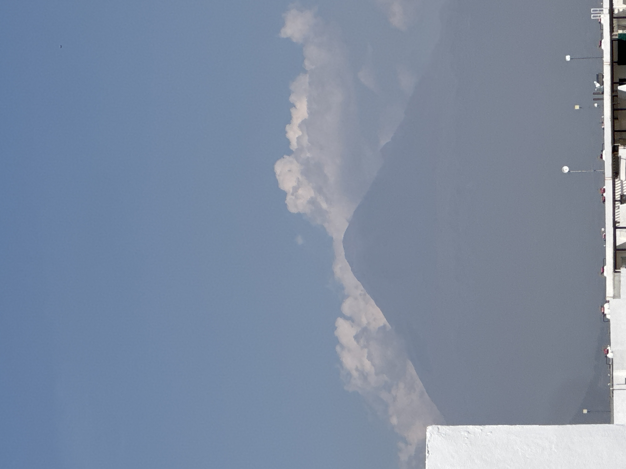The image presents a landscape near San Francisco Ocotlán, Mexico, capturing a blend of natural and man-made elements. A mountain range is prominent, partially obscured by clouds. To the right, a white building with multiple stories and street lights is visible. The sky is a clear blue, and there's a small bird in the top left corner.