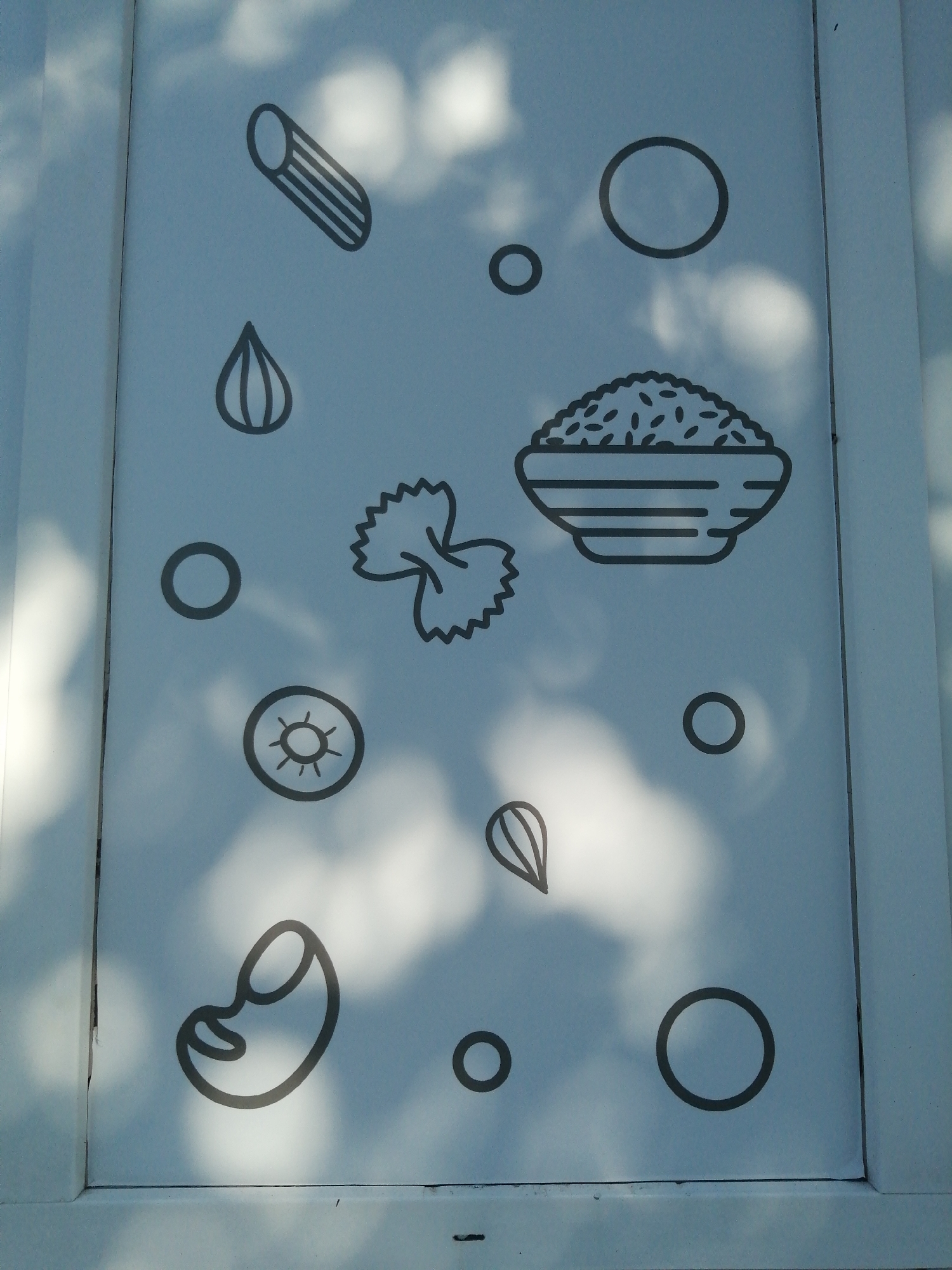 The image shows a light blue surface with several black line drawings of food items and circles. The food items include a penne pasta shape, a tear-drop shape that could represent a lentil or bean, a farfalle pasta shape, a bowl of rice, and a bean shape. There are also several plain circles scattered across the surface. The surface is illuminated by dappled sunlight, creating shadows and highlights that suggest it is outdoors, possibly on a panel or wall. The shadows appear to be from tree leaves. The image does not provide any explicit information about the city of Slonim, Belarus, or any specific context beyond the visual elements depicted.