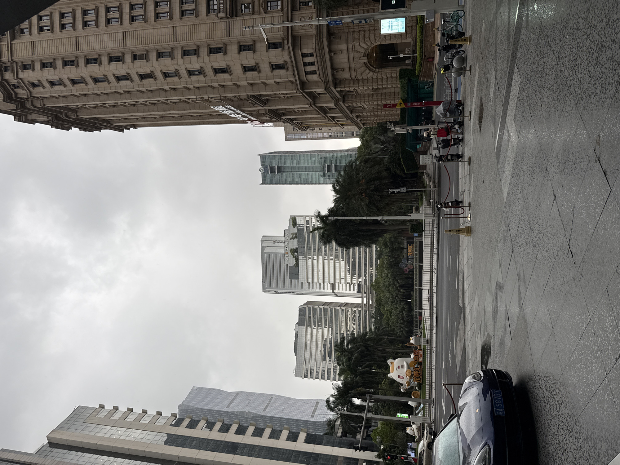 The media file depicts an overcast day in Tianhe District, China, likely in the morning or late afternoon. The scene is a city street with modern architecture. The main subjects are several tall buildings, some with glass facades reflecting the cloudy sky, and others appearing to be older, made of stone with numerous windows.
In the foreground, a dark blue car is parked on the right side of the street, its license plate partially visible as "A S8707". The street and sidewalk are wet, indicating recent rain, and the paving stones have a geometric pattern. Along the sidewalk, there are some decorative elements, including a large statue of a cat with coins, and smaller cat figures. A traffic light is visible in the lower left corner, showing red.
Further down the street, palm trees and other greenery are visible, interspersed between the buildings. There are several bicycles and electric scooters parked along a fenced-off area, suggesting a public transportation or rental system. Some people are visible walking and standing, though their features are not distinct.
The overall atmosphere is one of urban life on a cloudy day. Visible text includes "GRAND PLAZA" on one of the buildings and some Chinese characters on signs and the decorative elements, such as on the large cat statue. The sky is uniformly grey and cloudy, suggesting a lack of direct sunlight. The scene is static, capturing a moment in time on a typical day in this district.