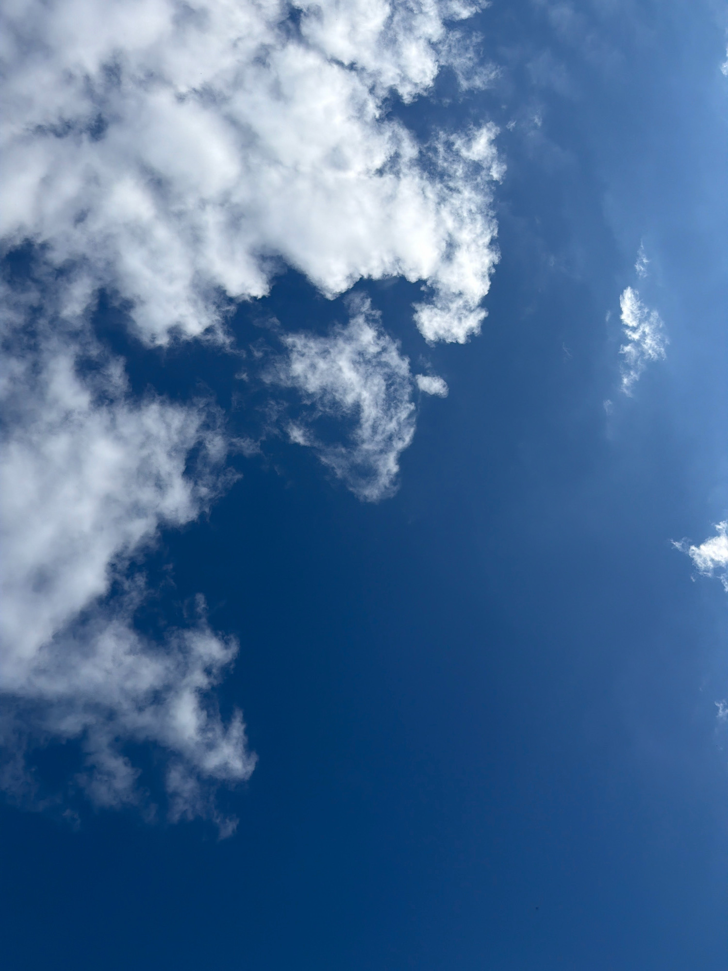 The image shows a vibrant, clear blue sky with scattered white clouds. The clouds are predominantly on the left side of the frame, with a large, fluffy formation taking up a significant portion of the upper left quadrant. Smaller, wispy clouds are dispersed throughout the rest of the sky. The image is captured from a low angle, looking directly upwards. There are no people, buildings, or any other discernible objects or activities visible in the scene, besides the sky and clouds. The location is identified as Бабініцкі сельскі Савет, Belarus.