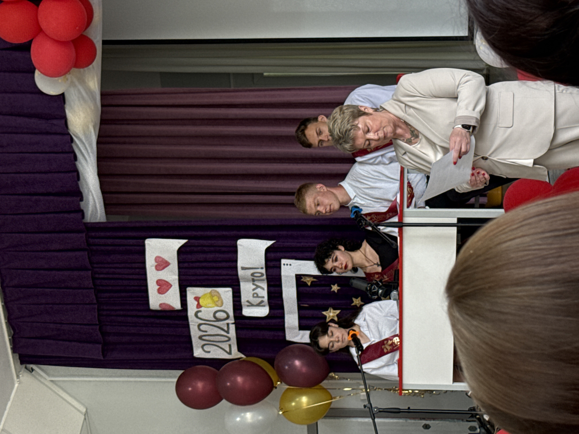 The scene depicts a group of people, possibly at an event, set against a purple backdrop. The backdrop is adorned with banners reading "2026" and "Круто!", along with decorative hearts and a bell. Balloons in red, white, and gold are scattered around the scene. At the forefront, a person in a light-colored suit is looking at a piece of paper. Behind them, three other people in white shirts with red sashes appear to be on a stage or platform, with microphones positioned near them. The overall setting suggests a celebratory or performance-based event. The location is noted as Unknown, Unknown.