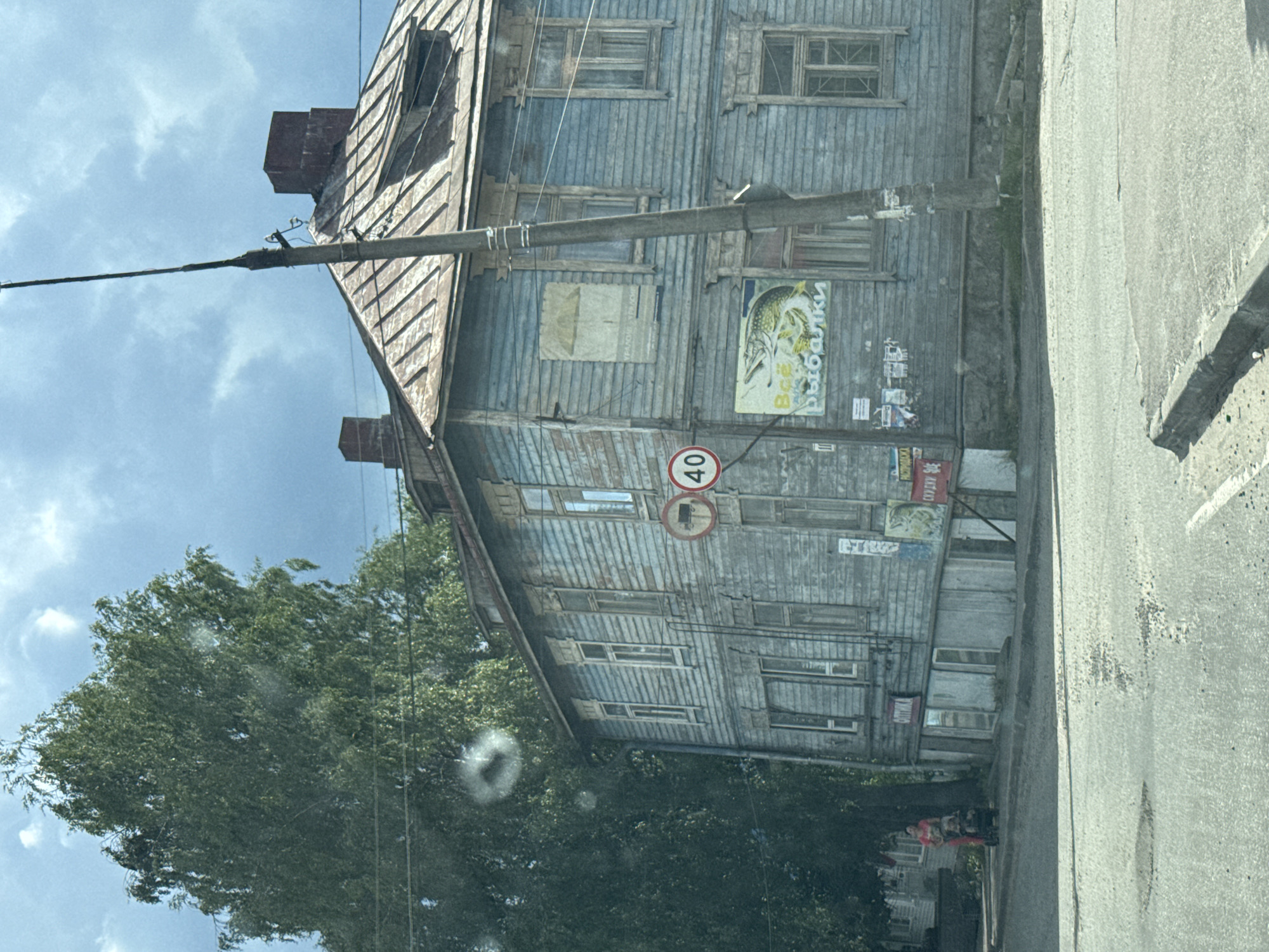 A weathered, light blue wooden building with multiple windows and visible repairs dominates the right side of the frame. The building has a metal roof with visible rust and a dormer window. A traffic sign indicating a speed limit of 40 is mounted on a pole to the left of the building. A large poster featuring a fish and Russian text is affixed to the lower portion of the building. A utility pole with wires runs vertically in front of the building. To the left, a large, leafy green tree stands tall. In the background, a cloudy sky with patches of blue is visible. In the lower left quadrant, a person in red clothing is pushing a stroller on the sidewalk next to the building. The foreground shows a paved road with some debris and cracks. The overall scene depicts a street view in a residential or commercial area.