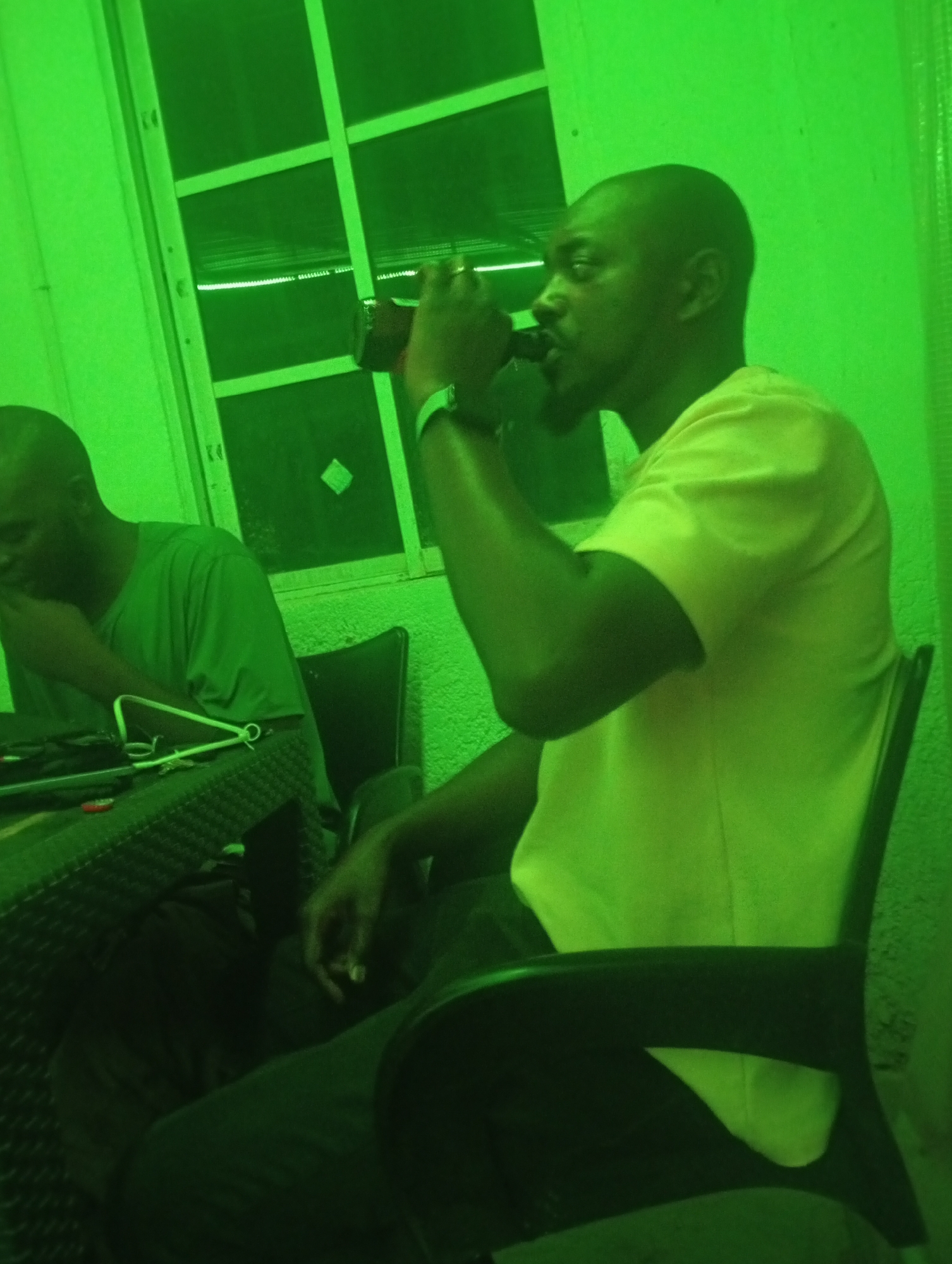 The image depicts two men in an indoor setting, heavily cast in a monochromatic green hue, suggesting either specific lighting or a night vision photograph. The location is Barakin Akawo, Nigeria.
In the foreground, a man with a shaved head and a short beard is seated in profile, facing left, actively drinking from a dark-colored can or bottle. He is wearing a light-colored, short-sleeved t-shirt, which appears light green due to the dominant lighting, and a watch is visible on his left wrist. He is seated on a chair with a dark frame.
In the background, to the left, another man with a similar physical appearance is seated at a table. He is looking down, seemingly engaged with an item on the table. He wears a darker green t-shirt. The table has a textured, possibly woven or rattan, surface. On the table, some wires or cables and a metallic, triangular object are visible.
Behind the men, a window with a prominent grid of panes is visible, looking out onto a dark area. The walls appear to be painted or rendered. The strong green tint obscures natural colors but contributes to a distinct, almost nocturnal, ambiance. No discernable text is visible on any objects or clothing. The overall scene conveys a casual, relaxed gathering.