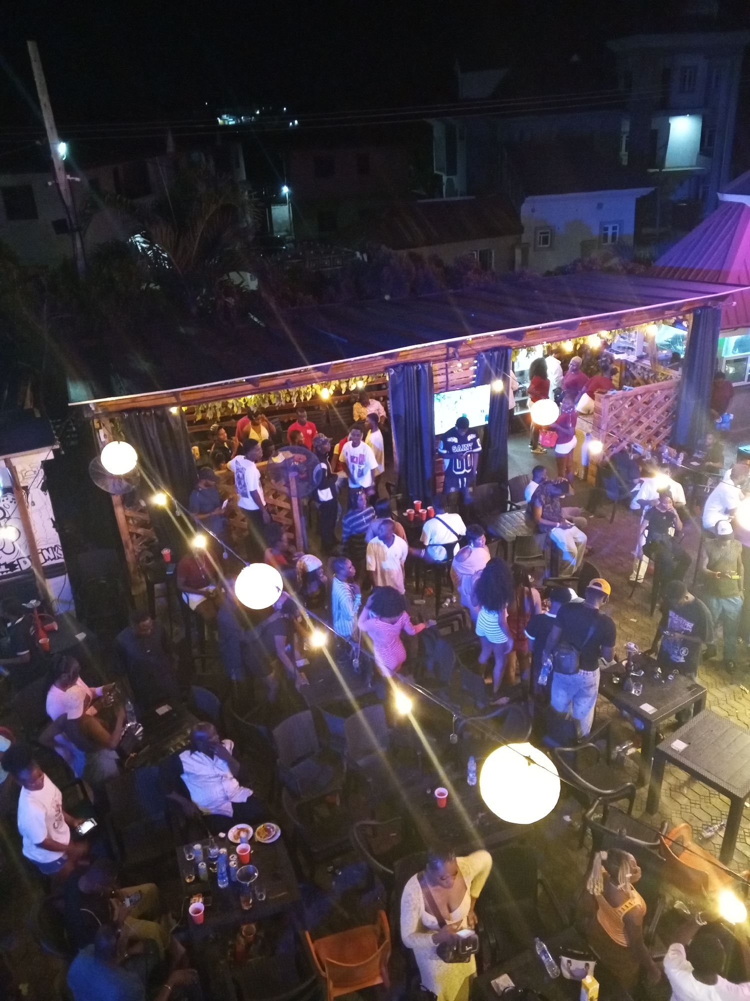 An outdoor bar in Lokoja, Nigeria, at night, viewed from above. A partially covered patio area is occupied by a crowd of people standing and sitting at tables. A television screen is mounted on a wall displaying an unidentified image. Overhead, round light fixtures illuminate the scene, with string lights hung loosely between them. Tables are scattered throughout, some with drinks and food. People are dressed in a variety of casual clothing. Buildings are visible in the background, indicating an urban setting.