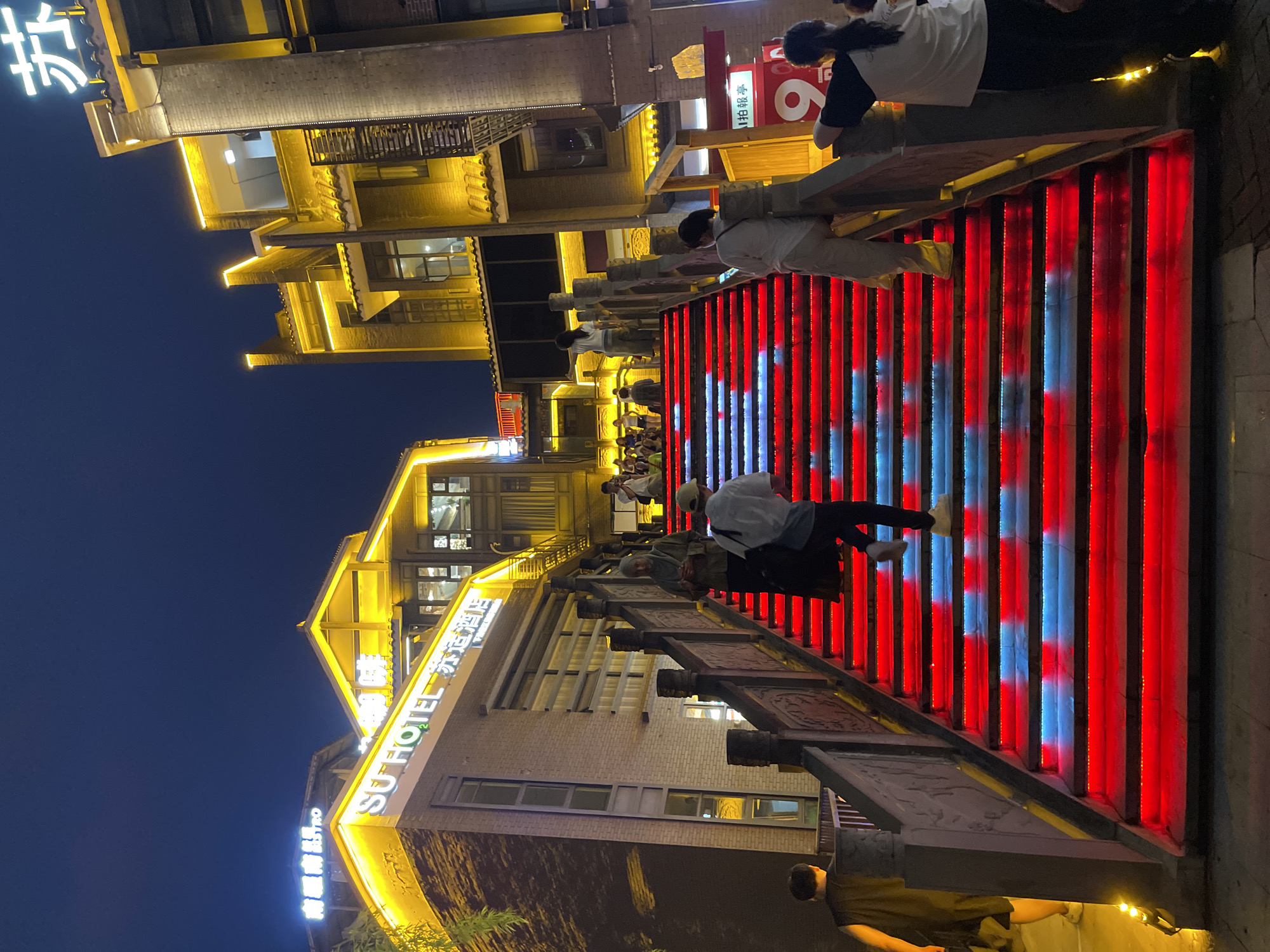 In Nan'an District, China, at night, a wide set of stairs is illuminated with vibrant red and blue lights, creating a striking visual effect. The stairs are bordered by ornate stone railings with carved details. Several people are ascending and descending the stairs, interacting with the environment. On the left, a person wearing a dark sleeveless shirt and shorts walks away from the camera. Further up the stairs, a woman in a headscarf and long clothing walks downwards, holding a phone. On the right side of the stairs, a person in a white shirt and beige pants is ascending. Another person in a black and white shirt and dark pants stands on the right side, looking towards the stairs. Above and to the left of the stairs, a building with yellow neon lights displays "SU HOTEL" and Chinese characters. Across from the hotel, another building with traditional Chinese architecture is also illuminated with yellow lights. The sky is dark, indicating nighttime. The overall scene depicts a lively urban environment with a focus on architectural lighting and pedestrian activity.